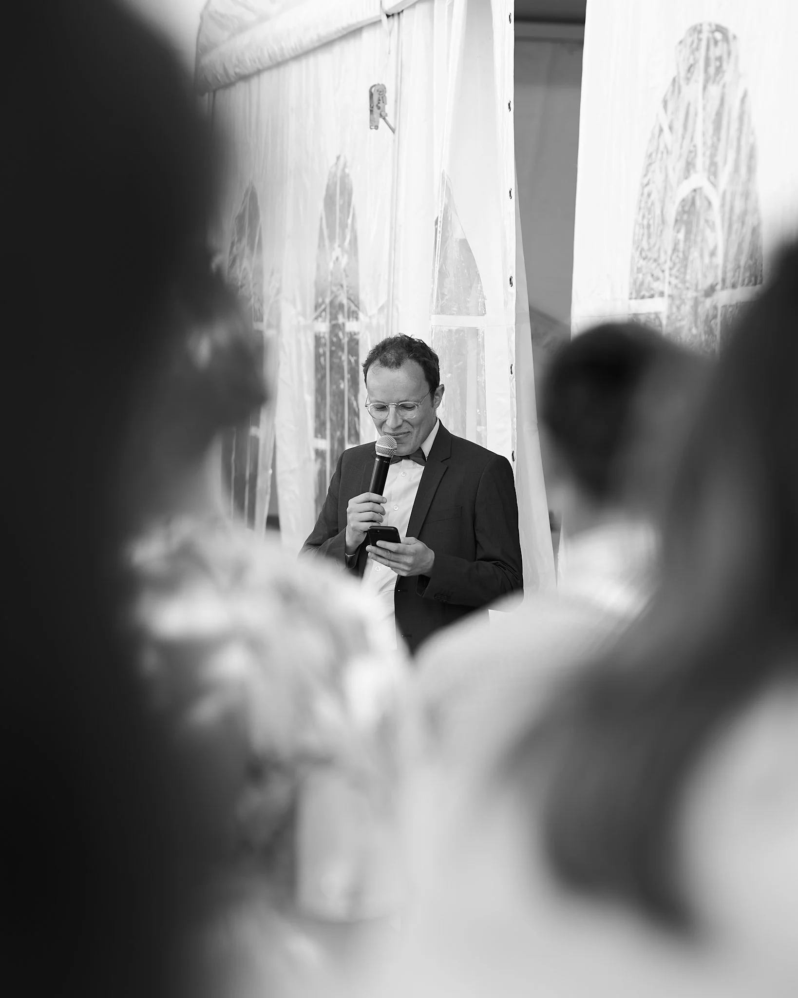Un homme en costume avec des lunettes parle ou lit un discours lors d'un événement, vu à travers une fenêtre, dans un contexte formel.