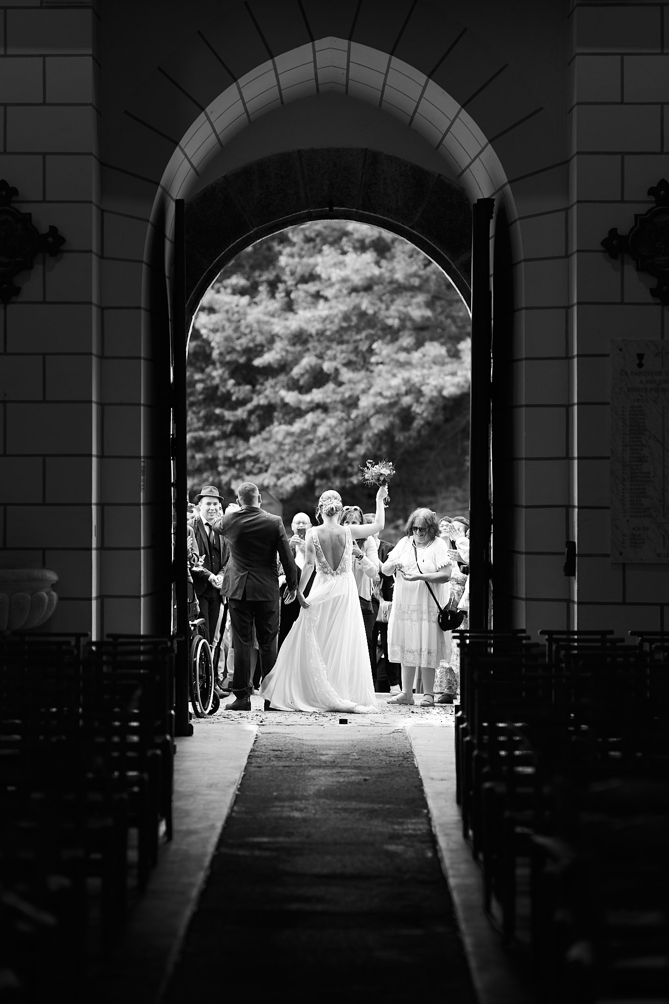 Mariage dans une église vue du fond, les invités accueillent la mariée en sortant à l'extérieur, scène en noir et blanc.