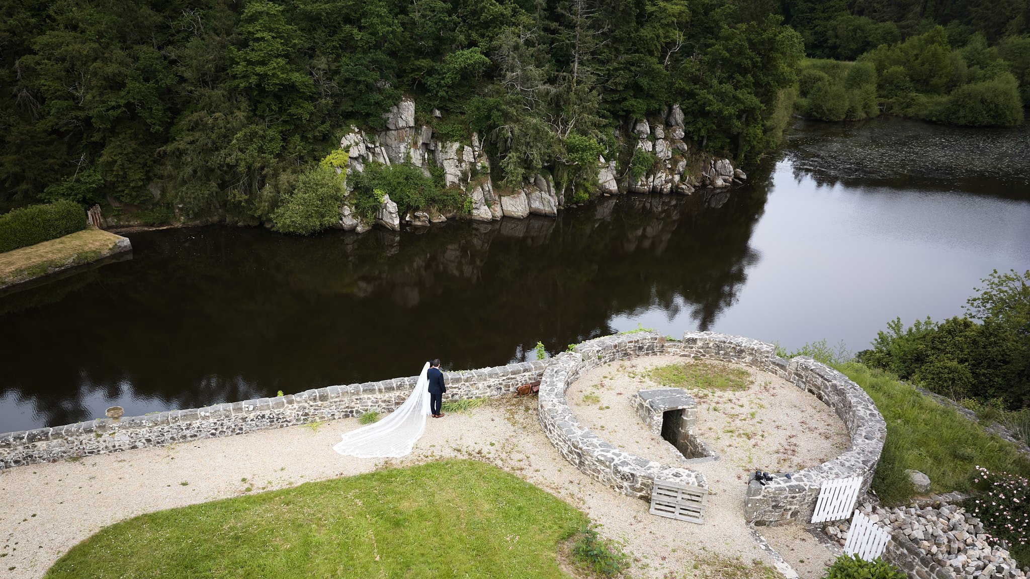 Un couple de mariés avec un voile blanc se tenant au bord d'une vieille ferme en pierre, surplombant une rivière calme entourée de verdure et de rochers.