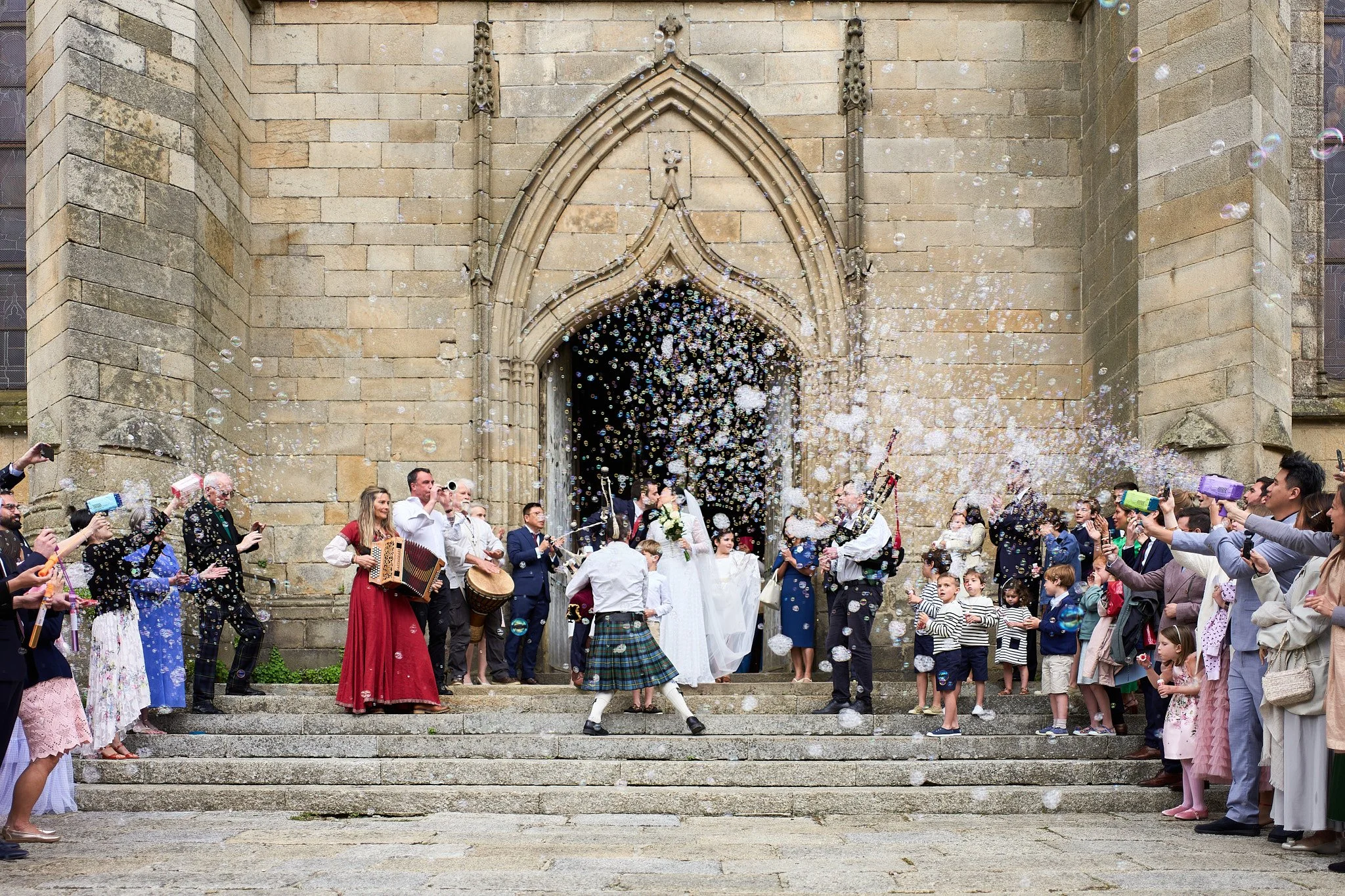 Célébration de mariage avec des invités lançant des bulles de savon devant une église en pierre.