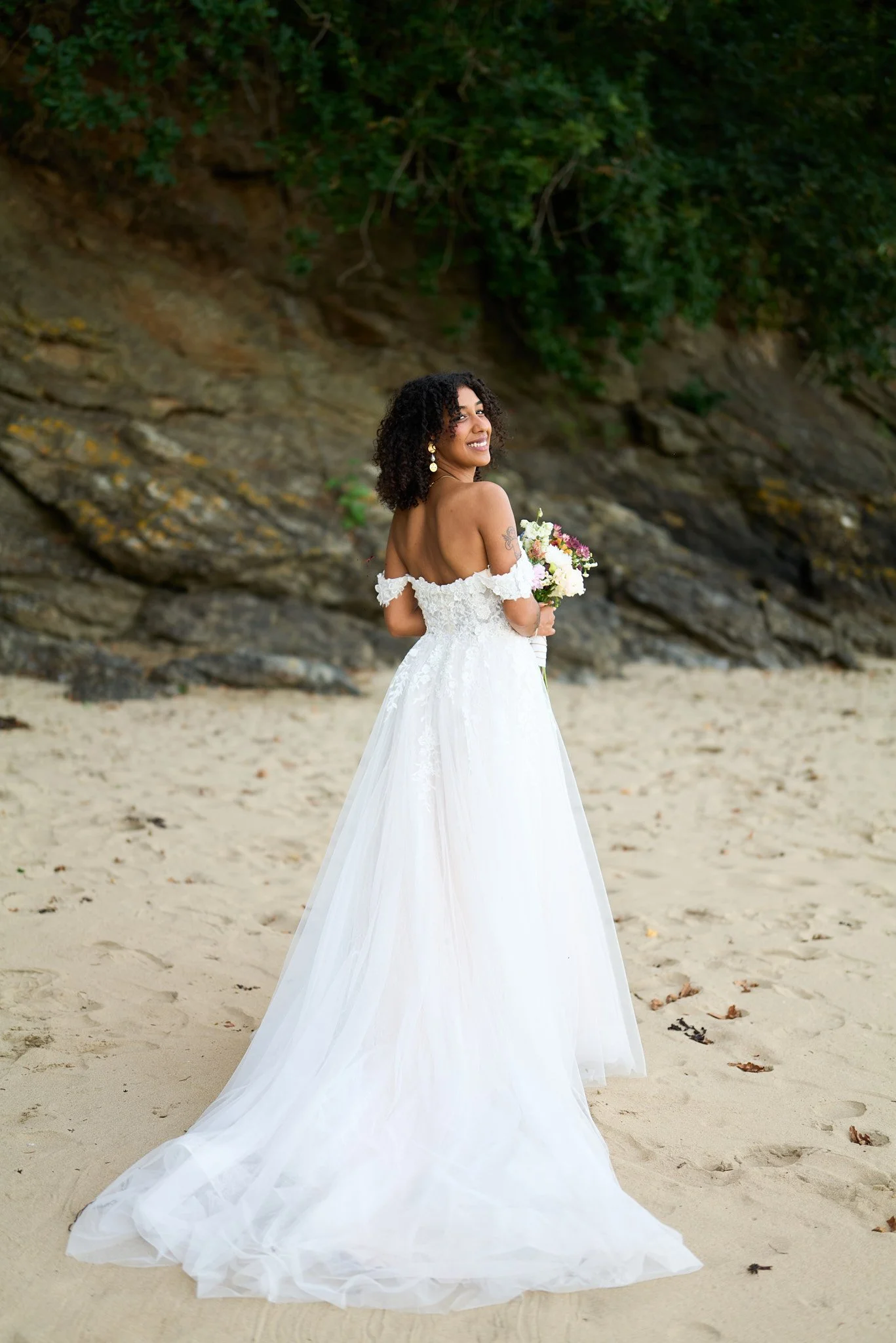 Femme en robe de mariage blanches, tenant un bouquet de fleurs, sur une plage, souriante, avec un rocher et des arbres en arrière-plan.