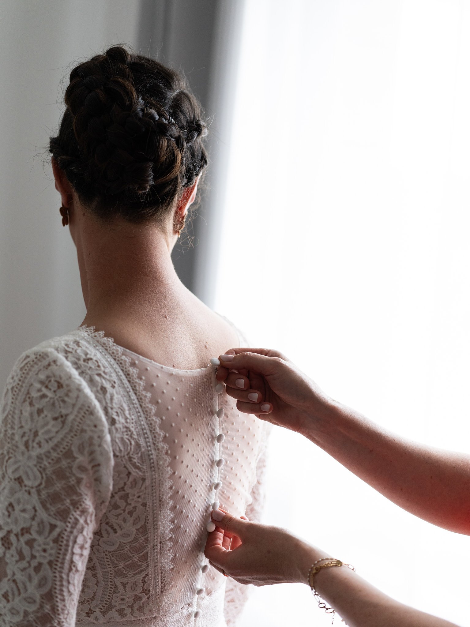 Une femme en robe de mariée à l'arrière, ses cheveux sont tressés. Une autre personne ferme les boutons du dos de la robe.