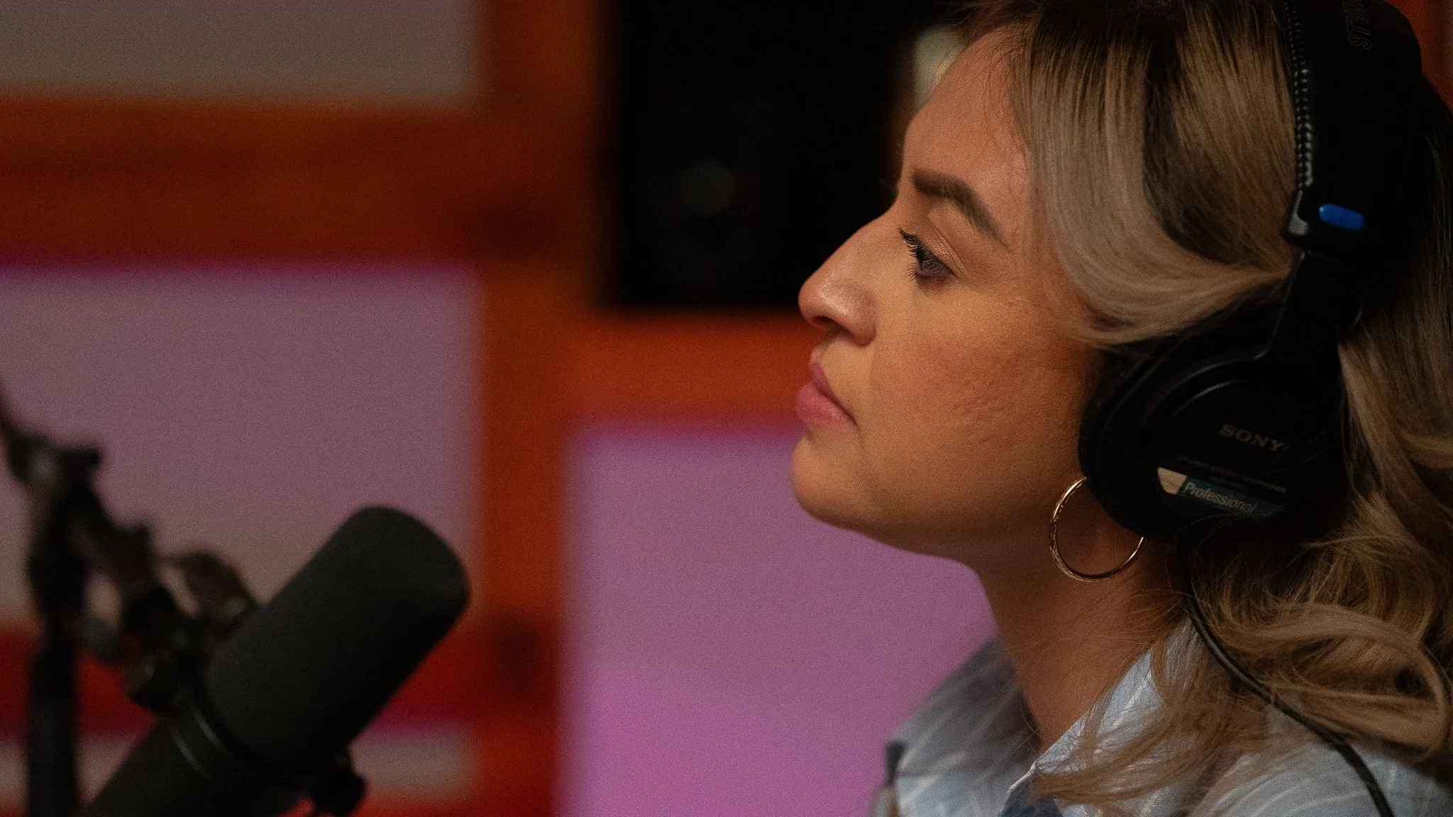 A woman wearing large black headphones sitting in front of a microphone in a recording studio.
