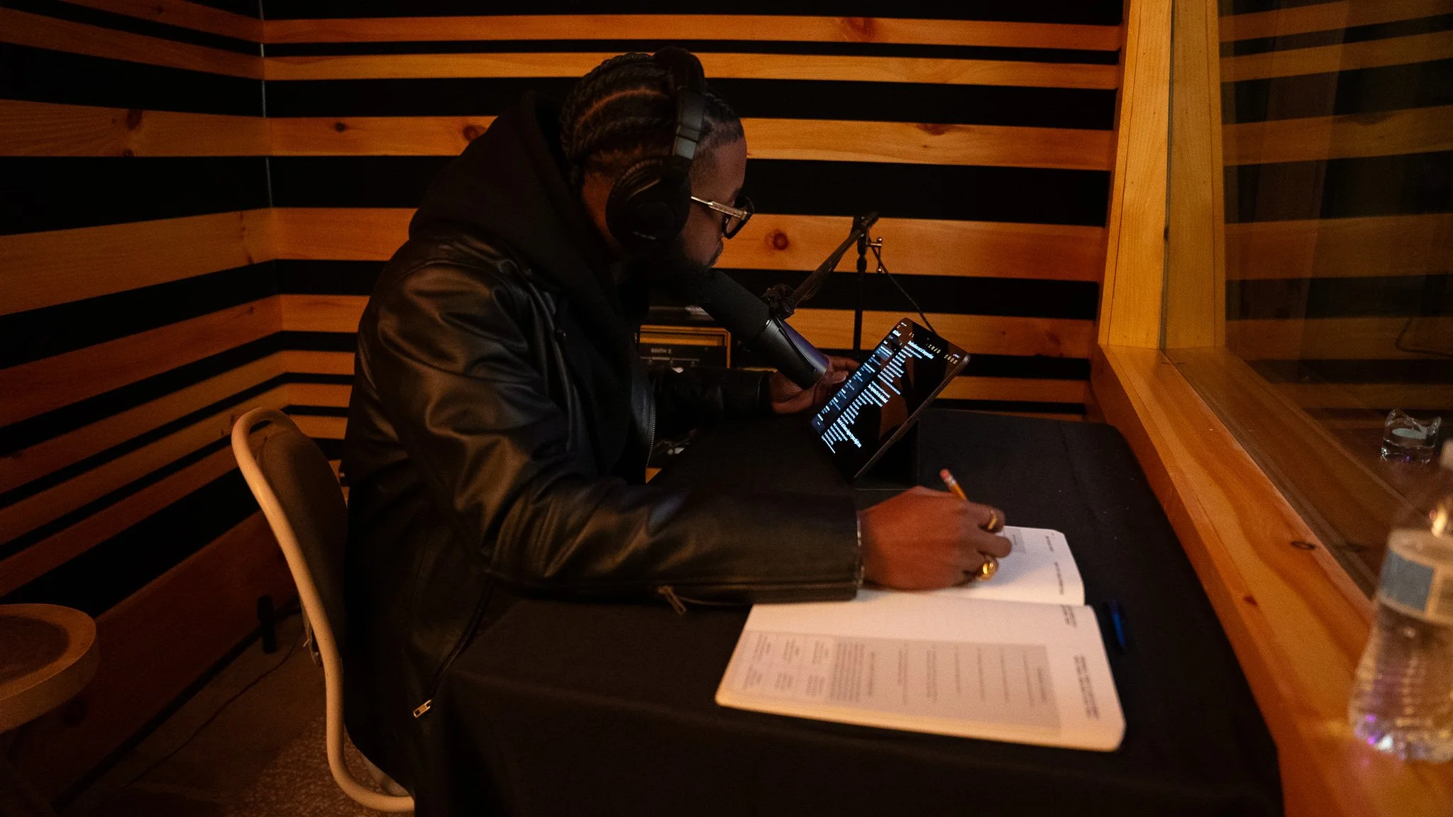 A man wearing glasses, headphones, and a black leather jacket is sitting at a table in a recording studio, reading a man with a notebook and a pen. There are bottles of water on the table. The studio has wooden acoustic panels.