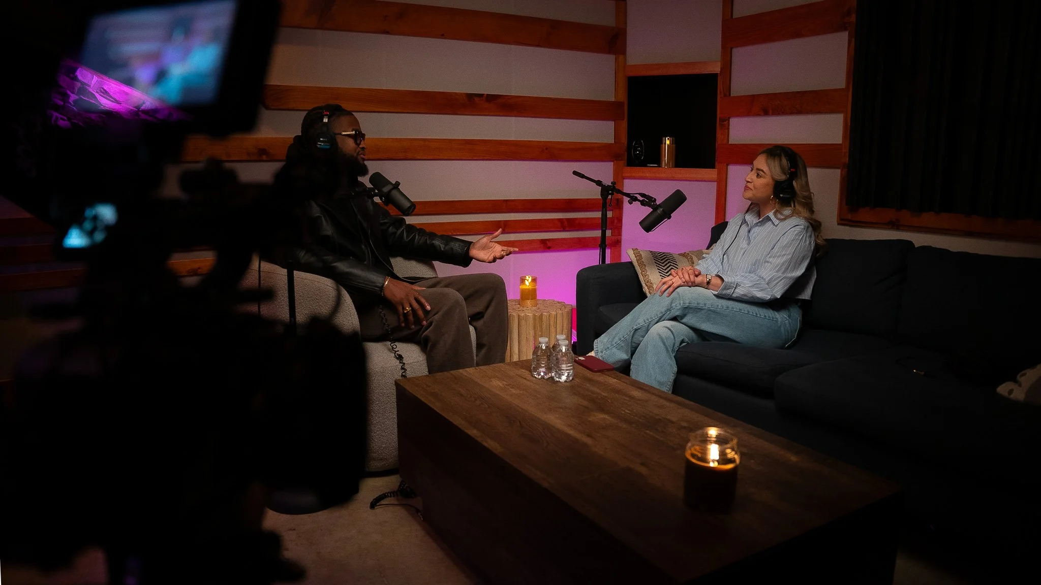 Two people recording a podcast in a cozy, dimly lit room with wooden wall accents and candles, some water bottles on the table, and microphones in front of them.