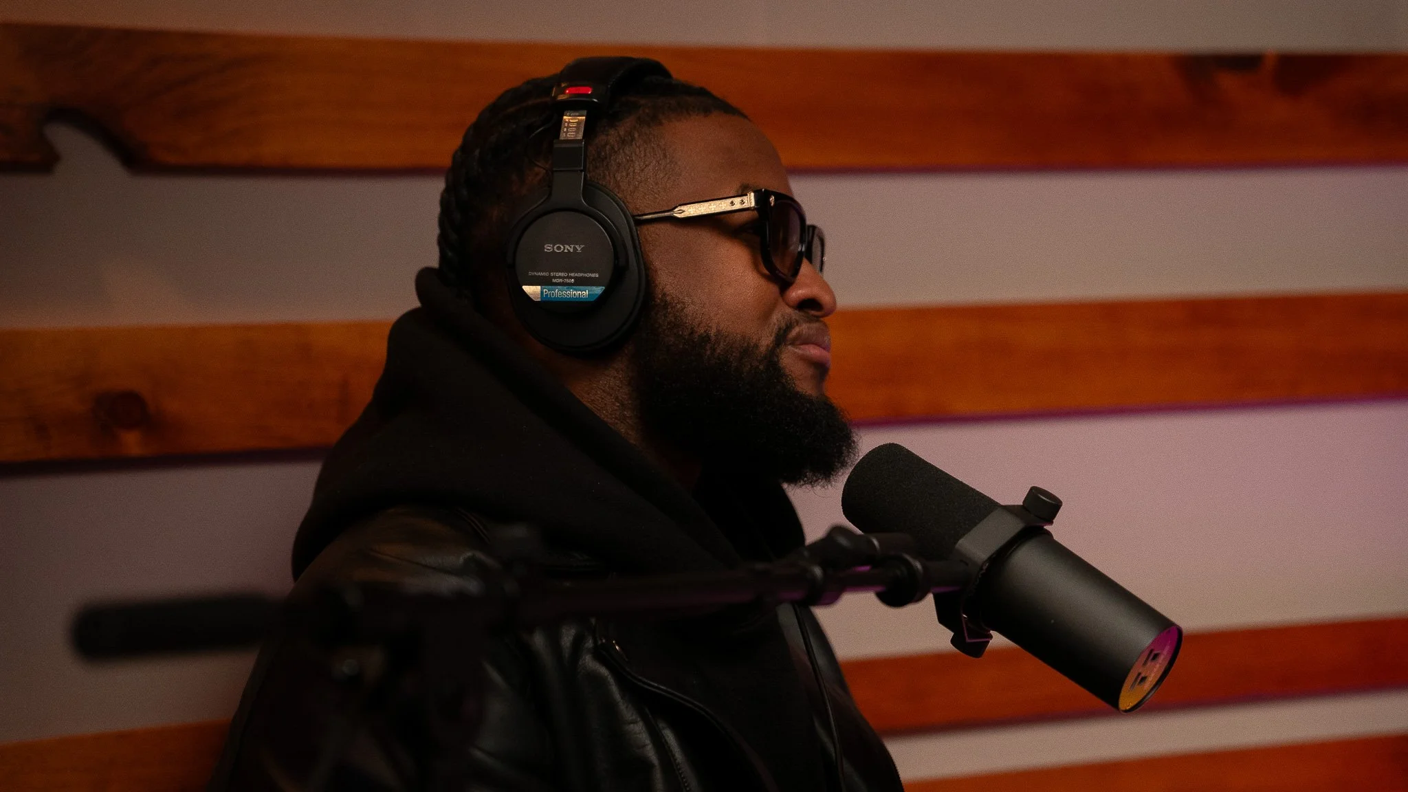 A man with a beard wearing sunglasses and Sony headphones is speaking into a microphone in a recording studio with wooden wall panels.