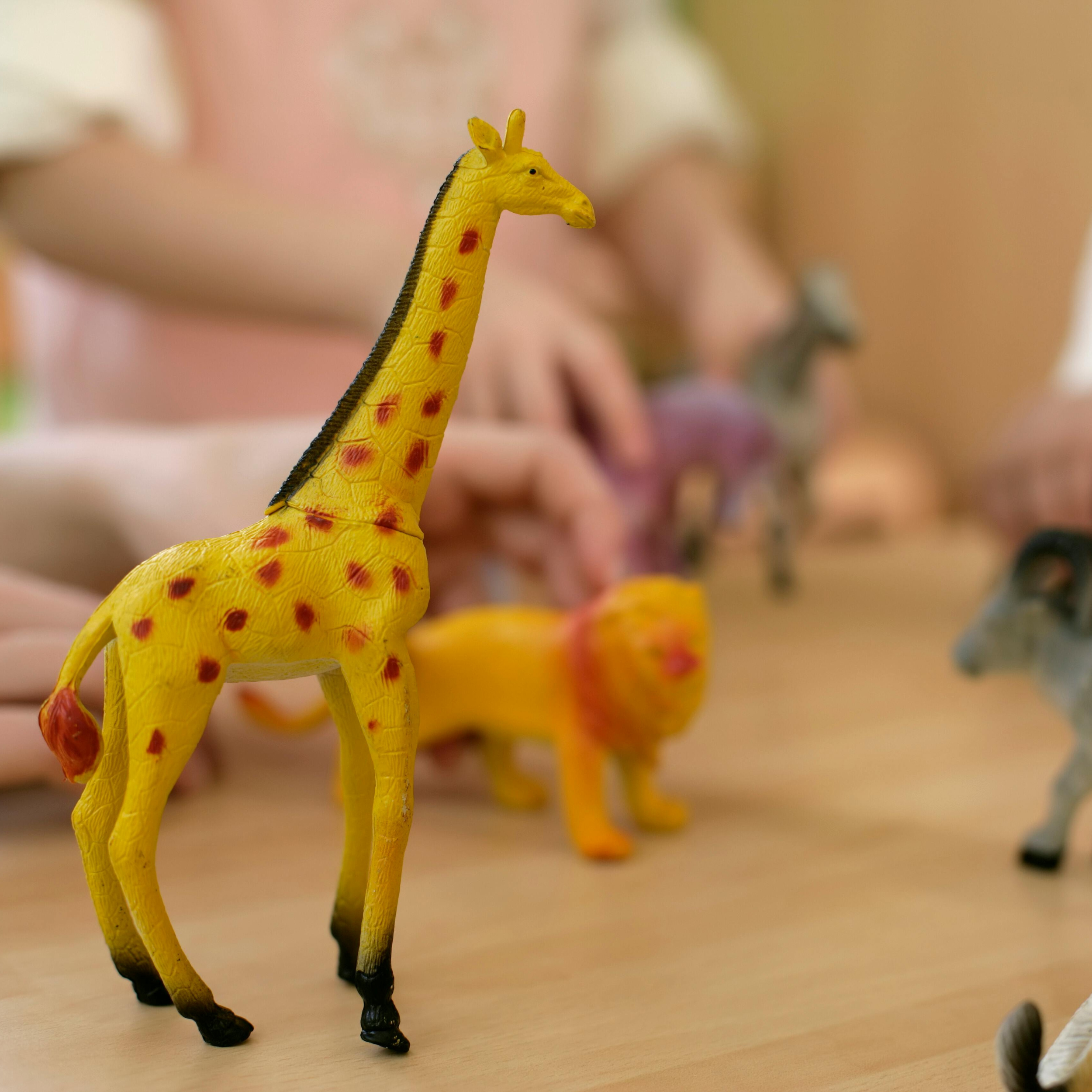 A collection of plastic toy animals on a wooden surface, including a yellow giraffe with orange spots in the foreground.