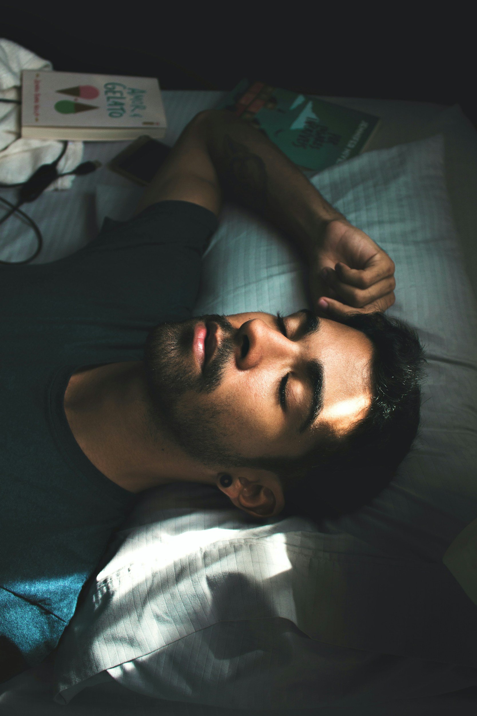 A man with closed eyes sleeping on his back in bed with sunlight casting shadows on his face, surrounded by books, a phone, and a charging cable.