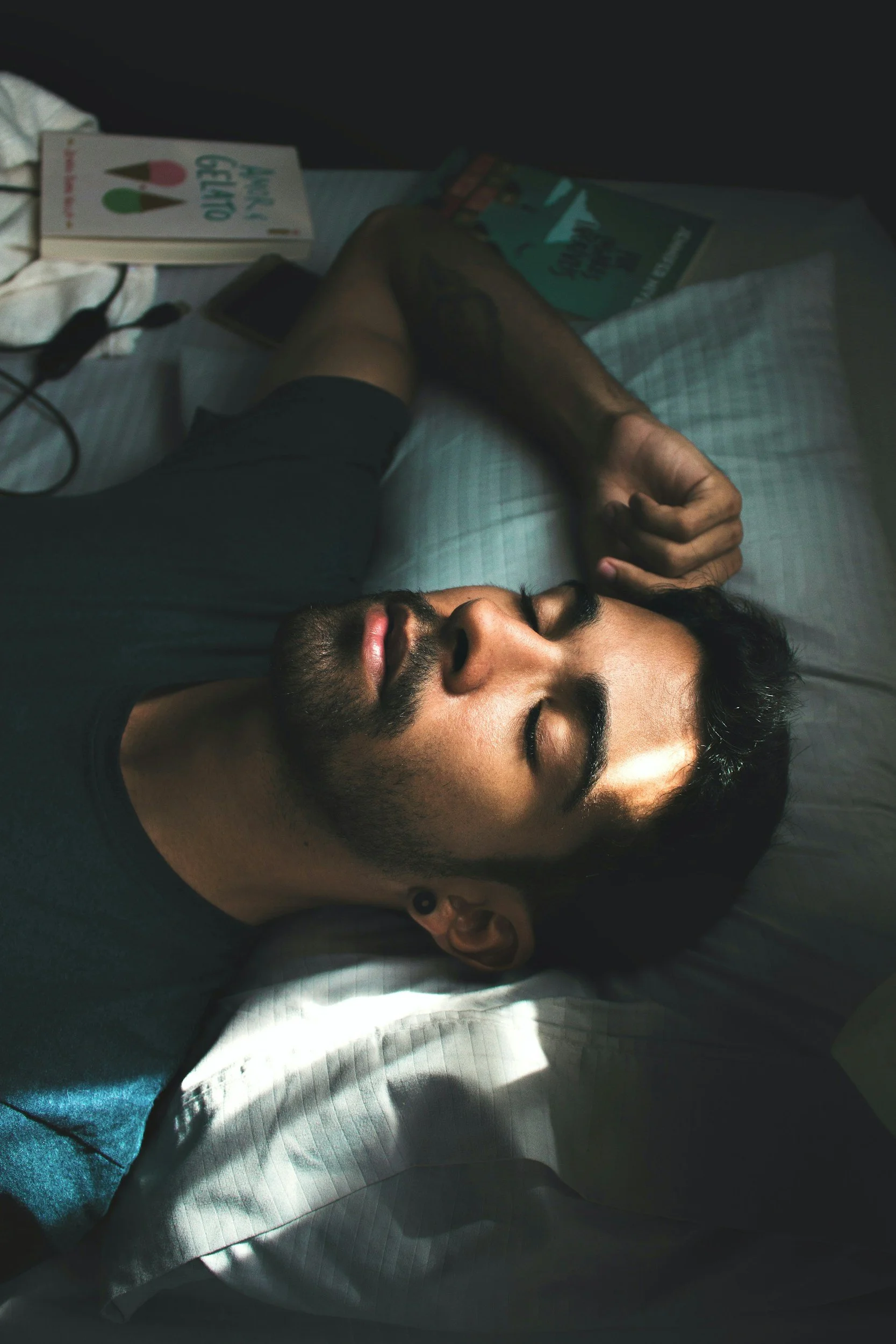 A man sleeping on a bed with his eyes closed, his right arm resting behind his head, sunlight casting shadows on his face, with books and a phone on the bedside table.