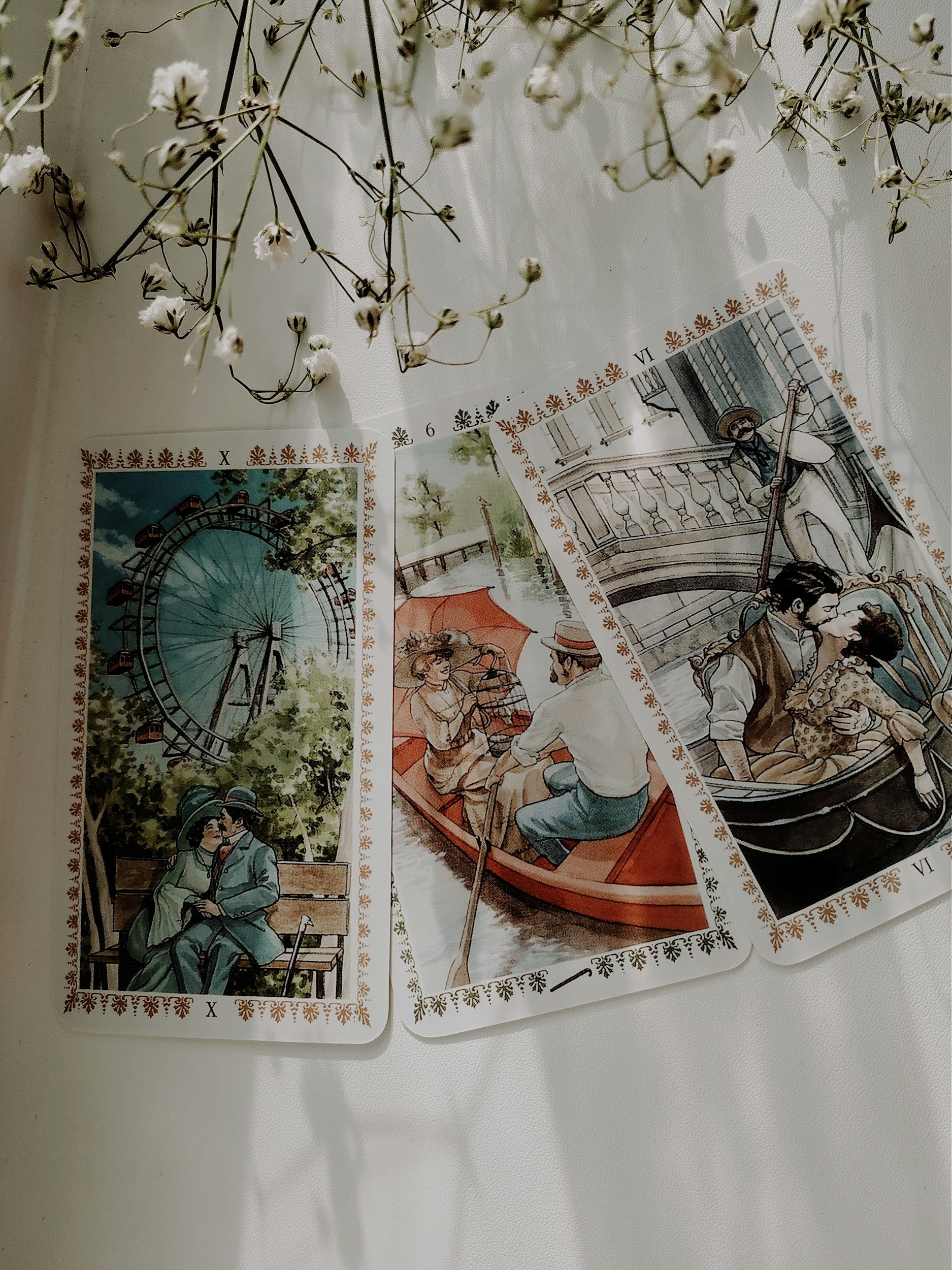 Three vintage-style tarot cards laid out on a white surface with dried white flowers above them. The left card depicts a couple sitting on a park bench with a Ferris wheel in the background. The middle card shows a woman and a man in a rowboat on a canal, the woman holding an umbrella. The right card features a romantic scene of a man and woman in a gondola, with a man playing a guitar in the background.