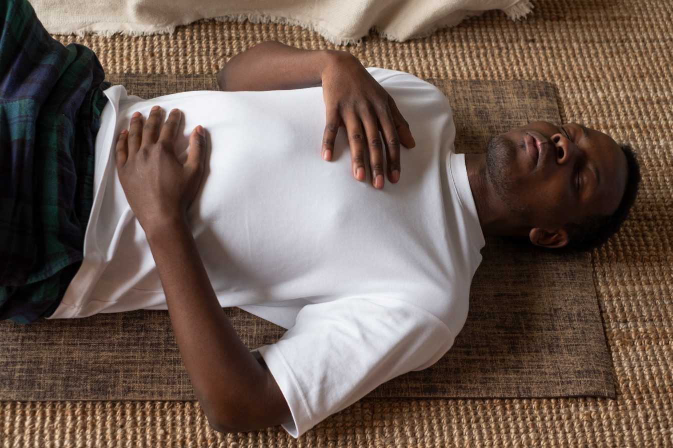 A man lying on his back on a yoga mat with eyes closed, resting, with hands on his chest, on a woven rug.