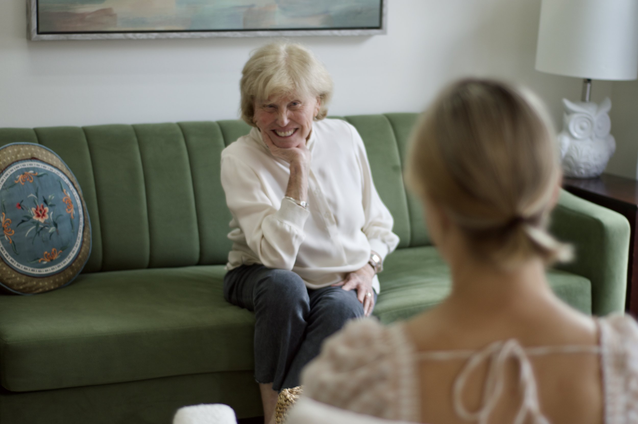 Two women having a conversation in a cozy living room, sitting on a red couch and beige armchair, with a decorative mirror and framed pictures on the wall.