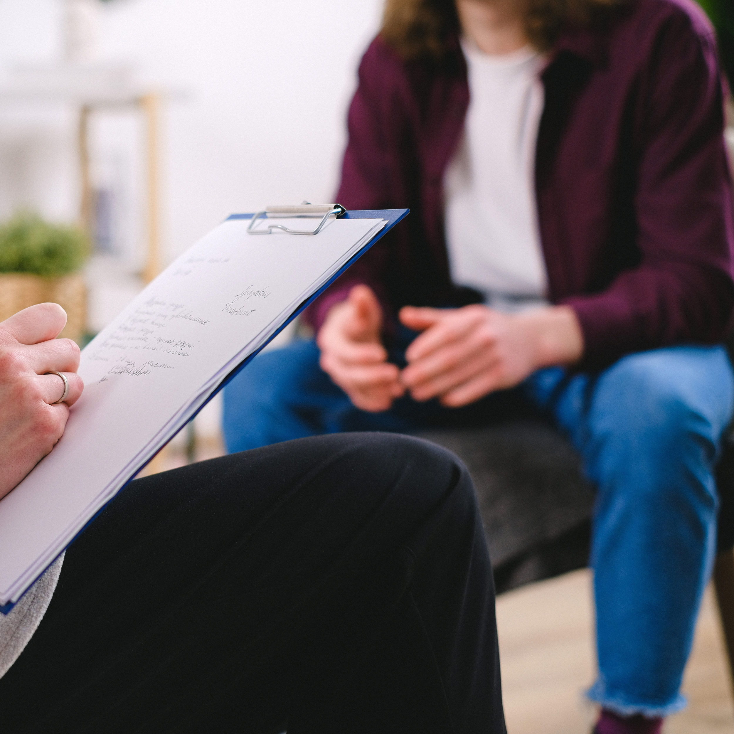 A person in a therapy session holding a clipboard with notes, sitting in front of another person who is seated and listening attentively.