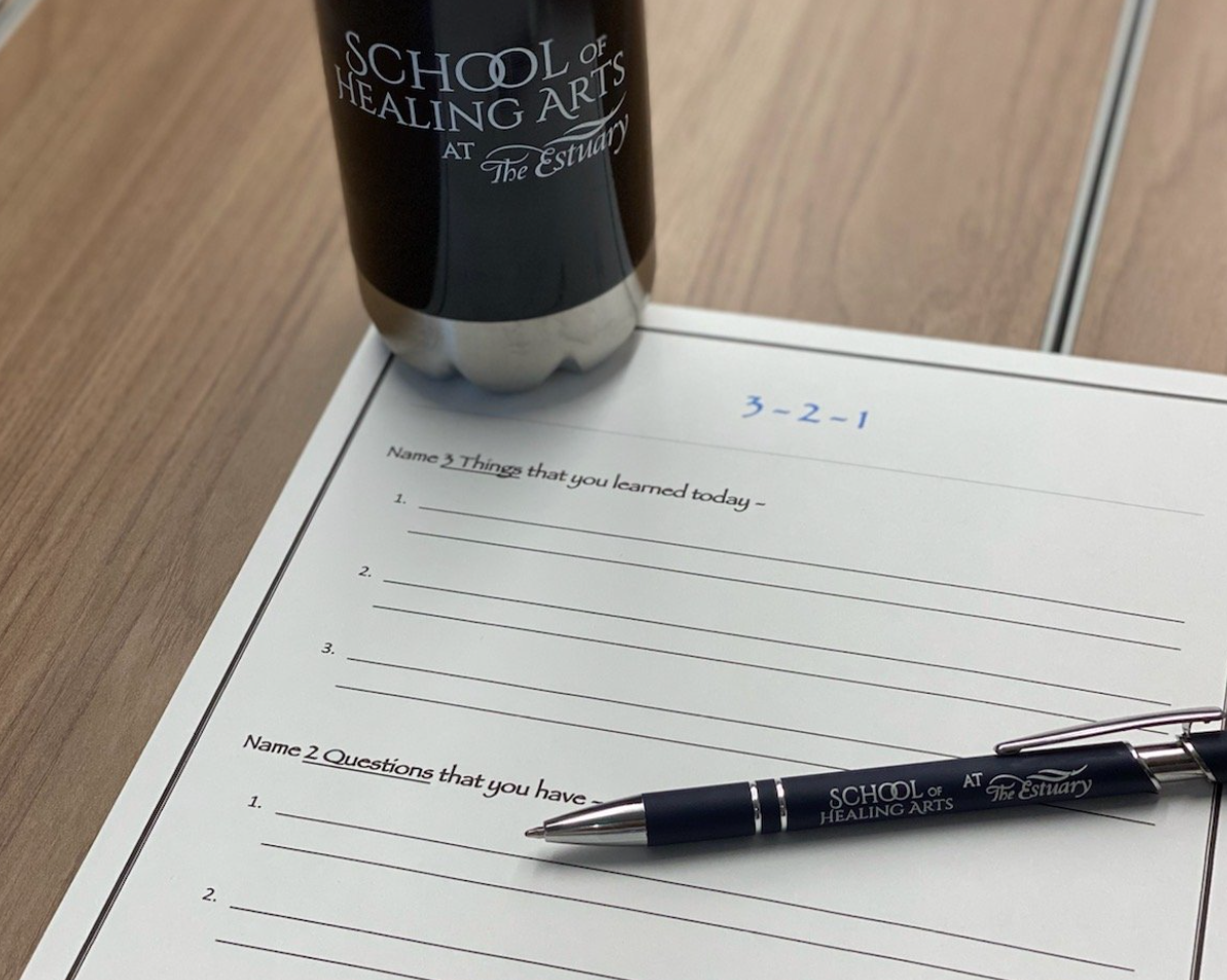A black pen with the words "School of Healing Arts at The Estuary" written on it, resting on a worksheet with the same text. The worksheet is titled "Name 2 Things that you learned today" and has numbered lines for answers. A water bottle with similar branding is placed on the worksheet on a wooden table.
