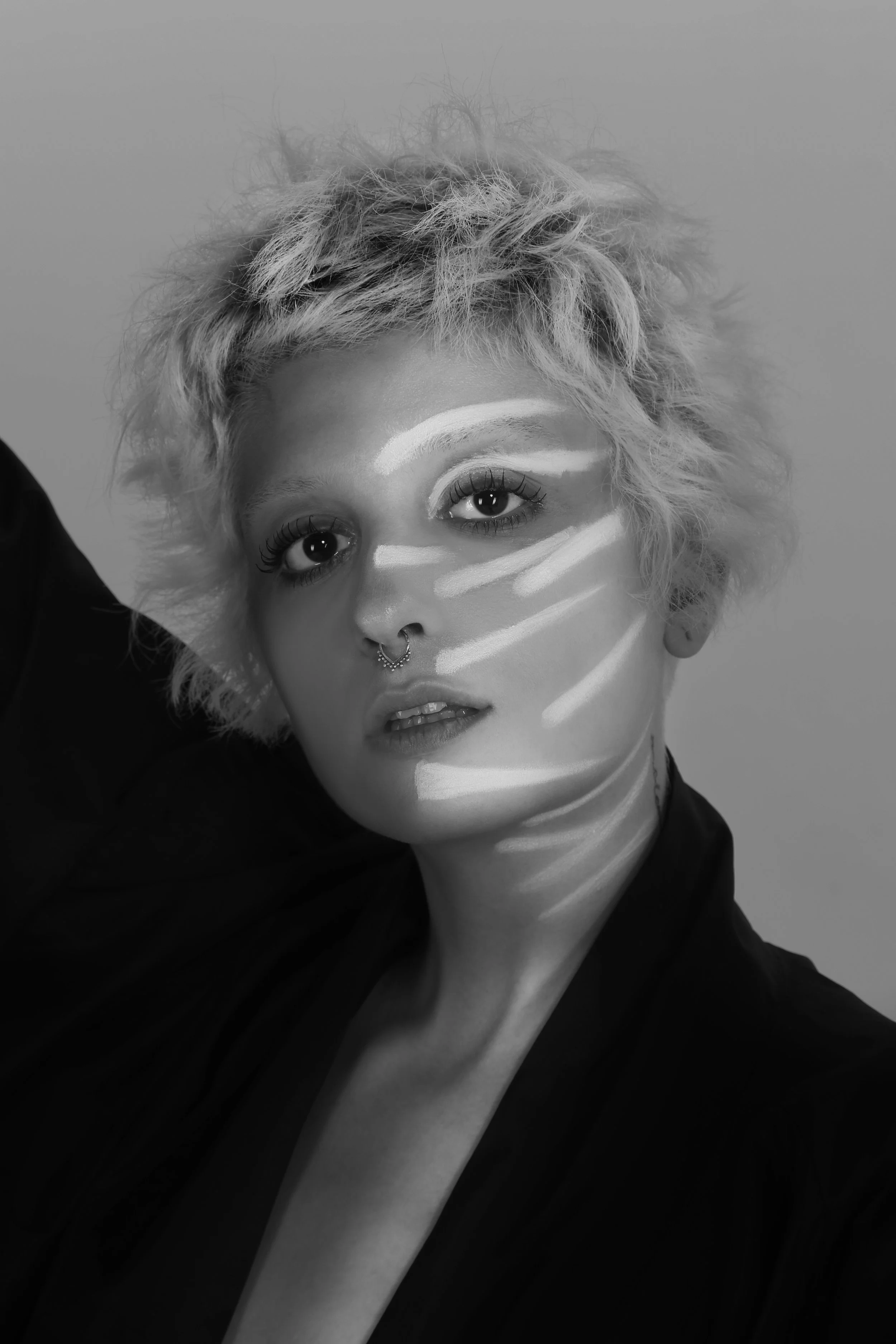 Black and white portrait of a woman with curly, short blonde hair, makeup, and a septum piercing, with light streaks on her face, posing with one arm raised.