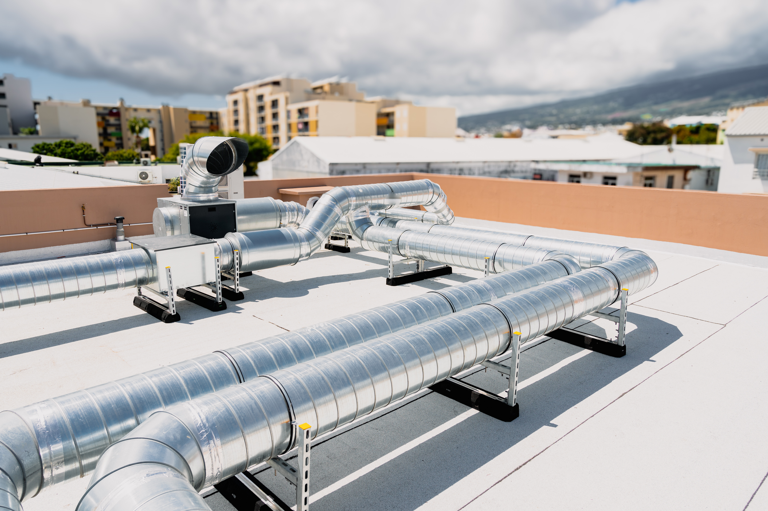 Multiple large silver ventilation ducts and exhaust pipes on a rooftop against a cloudy sky, with residential buildings and mountains in the background.