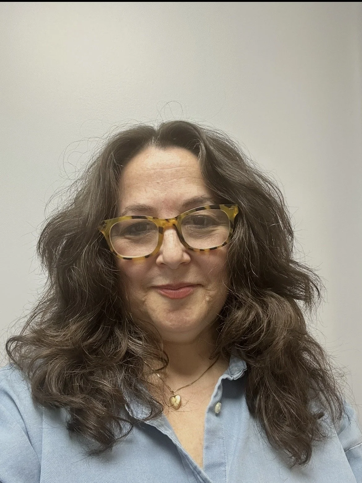 Close-up of a woman with brown hair, brown eyes, wearing tortoise shell glasses and a blue top, standing against a grey background.