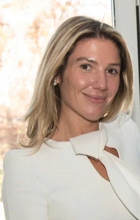A smiling blonde woman with earrings, wearing a blue floral shirt, indoors with a window in the background.