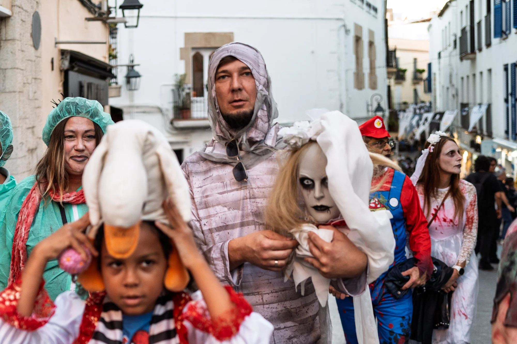 Man in bandages carries a veiled doll's head through the crowd, a child in a duck hat in front, Sitges — Leica Q3