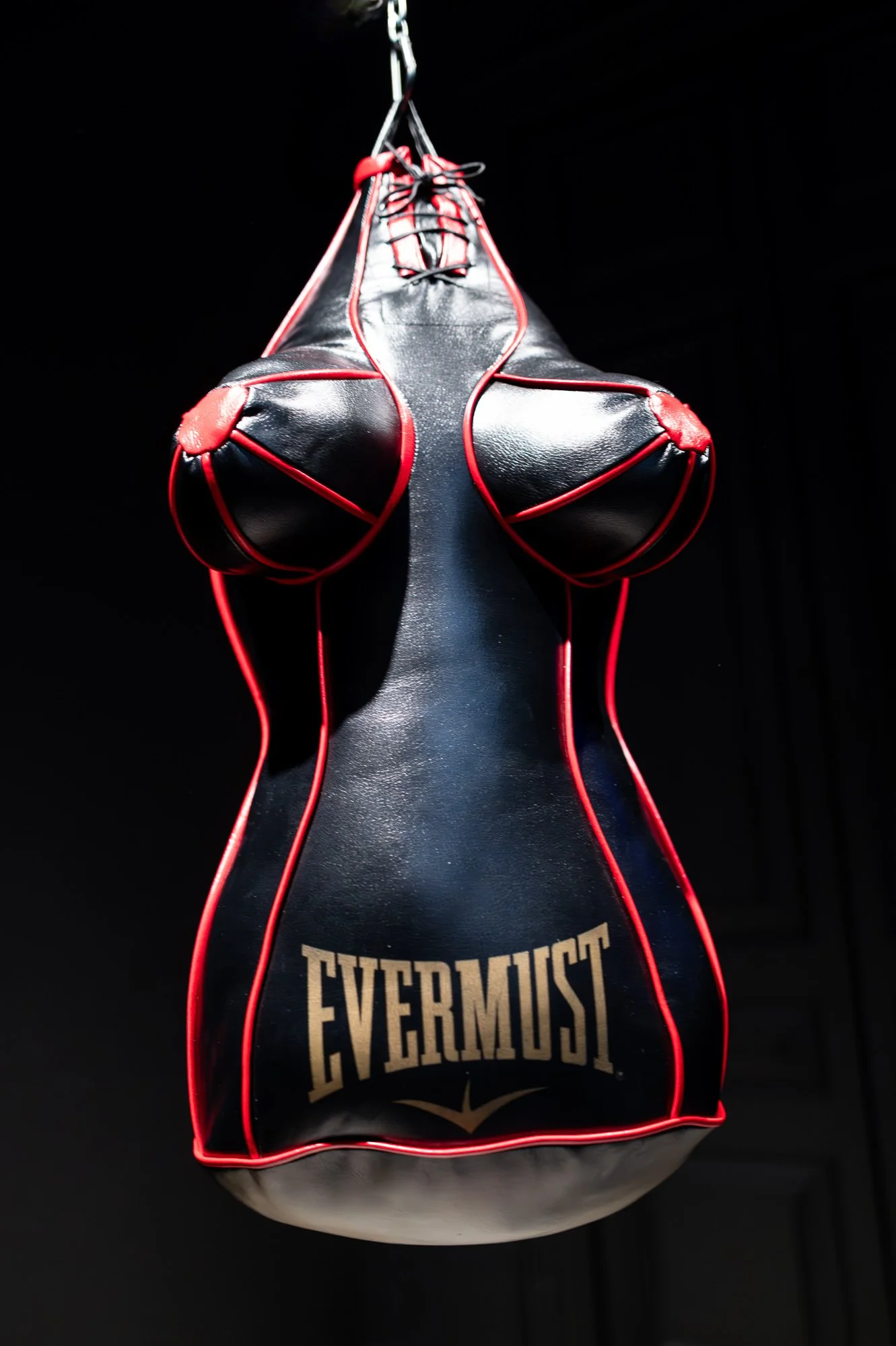 A black leather punching bag shaped like a woman's torso, with red piping and a gold logo, hanging against a black background