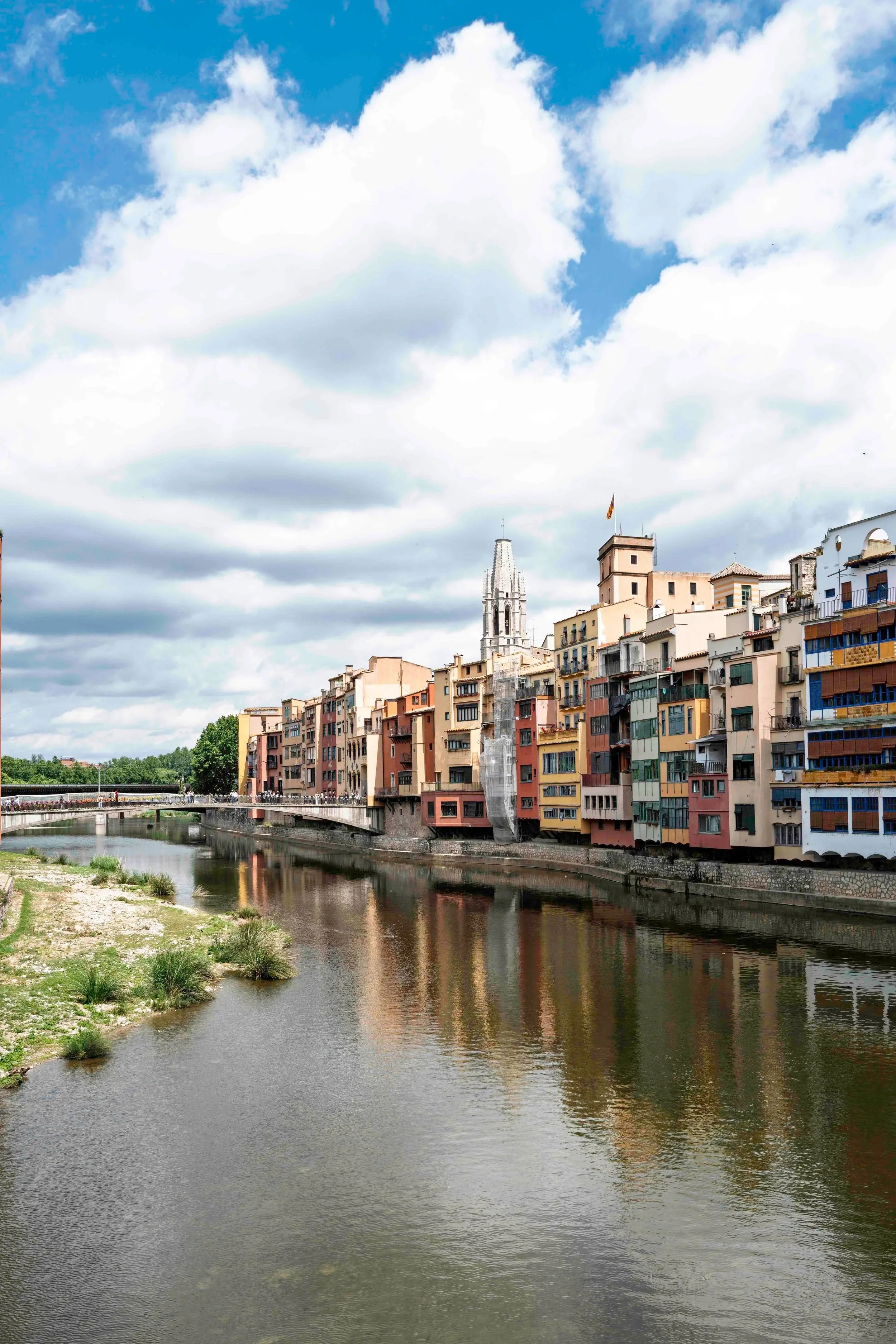 View along the Onyar River with rows of colourful historic buildings reflected in the water and the Cathedral tower in the background, Girona — Leica Q3
