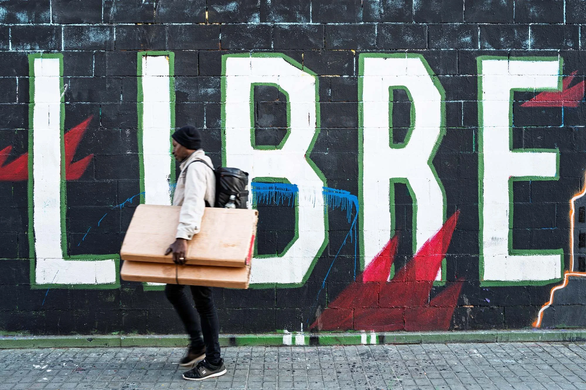 Man carrying wooden boards past a "LIBRE" graffiti mural at the Tres Xemeneies, Barcelona street art — Leica Q3