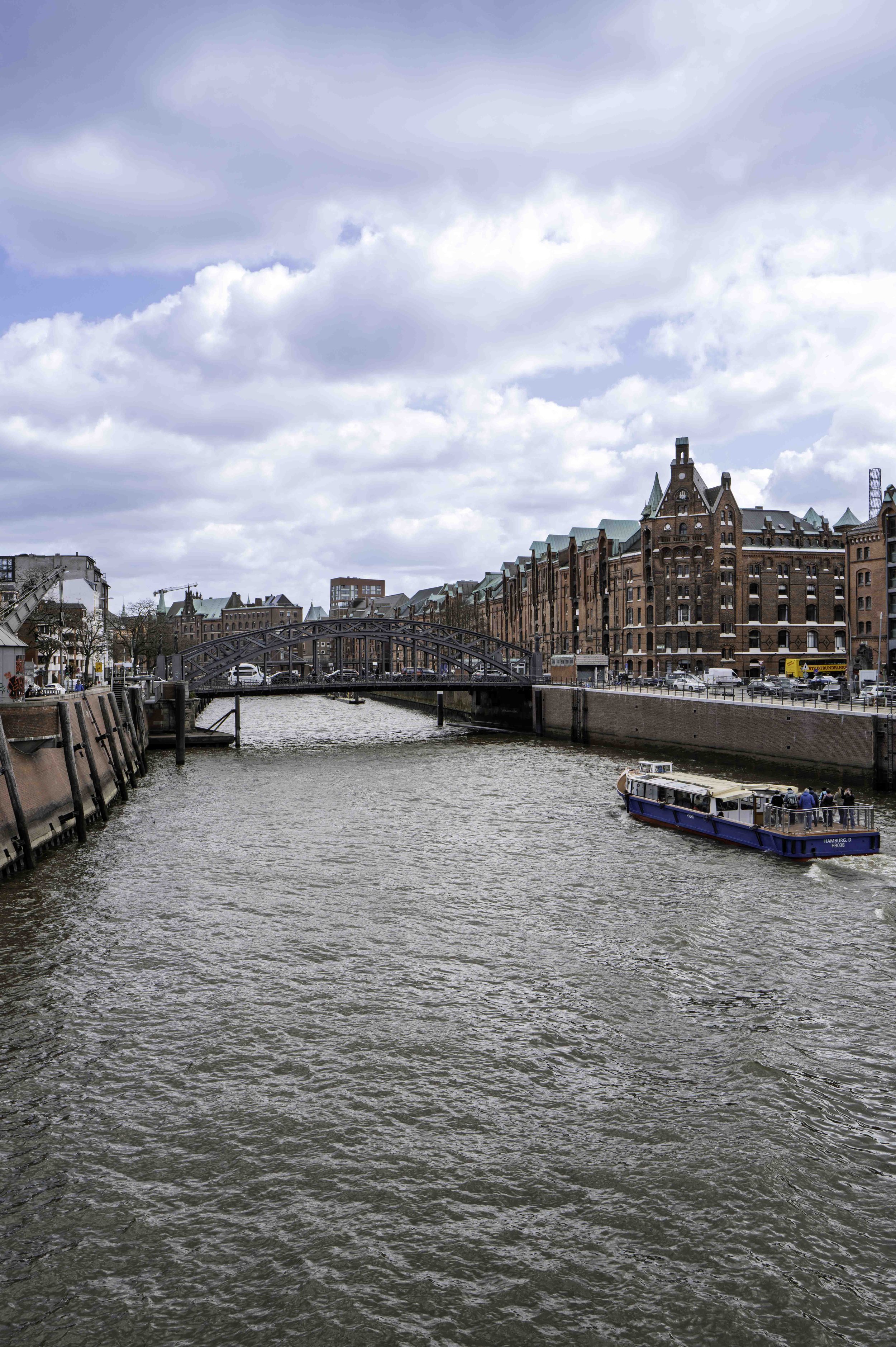 Blue harbour ferry passing under iron bridge on Hamburg canal, Speicherstadt warehouses in background — Leica Q3