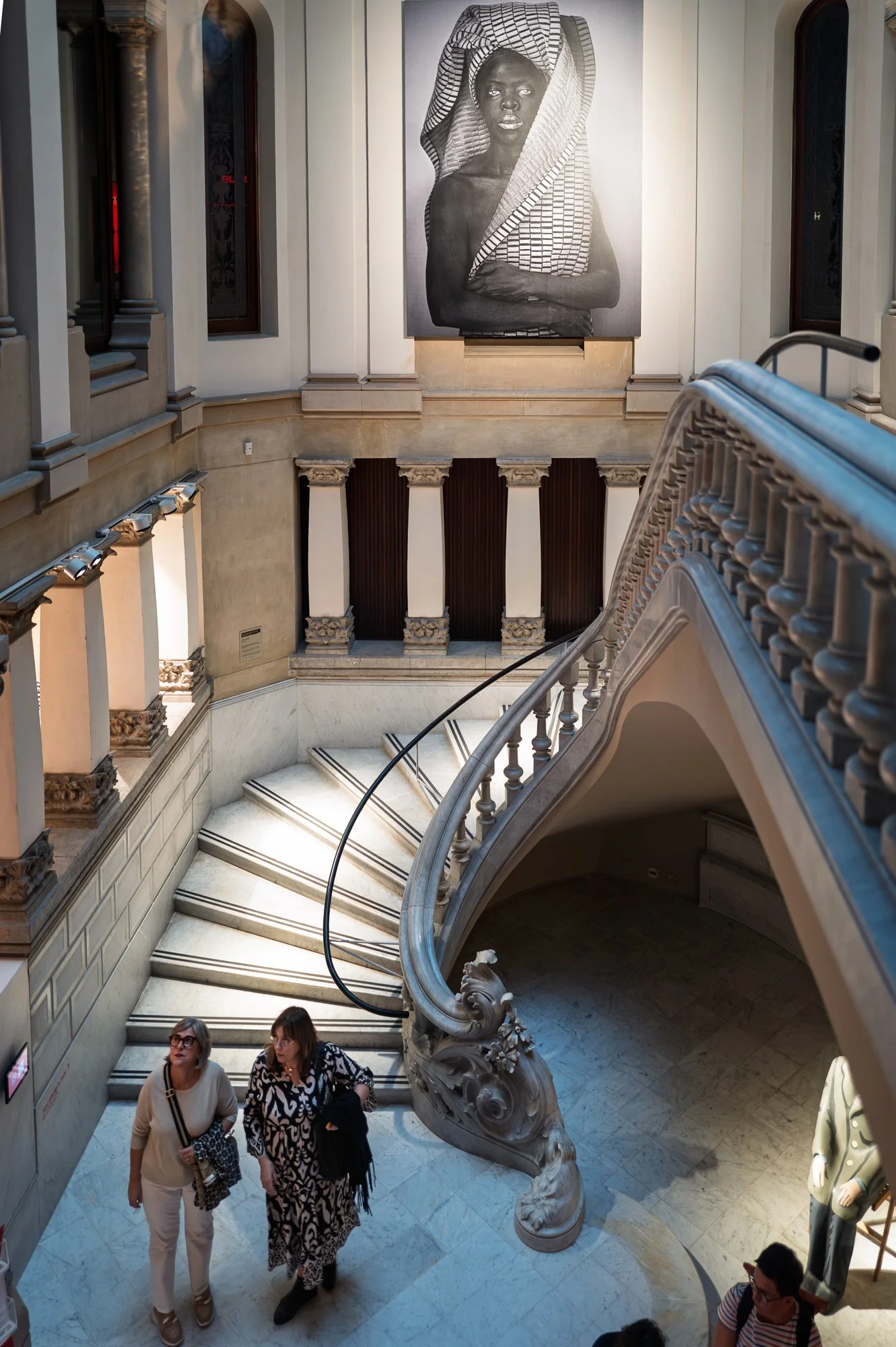 Two women at the foot of the marble staircase look up at the portrait above them, a carved newel post spiralling beside them