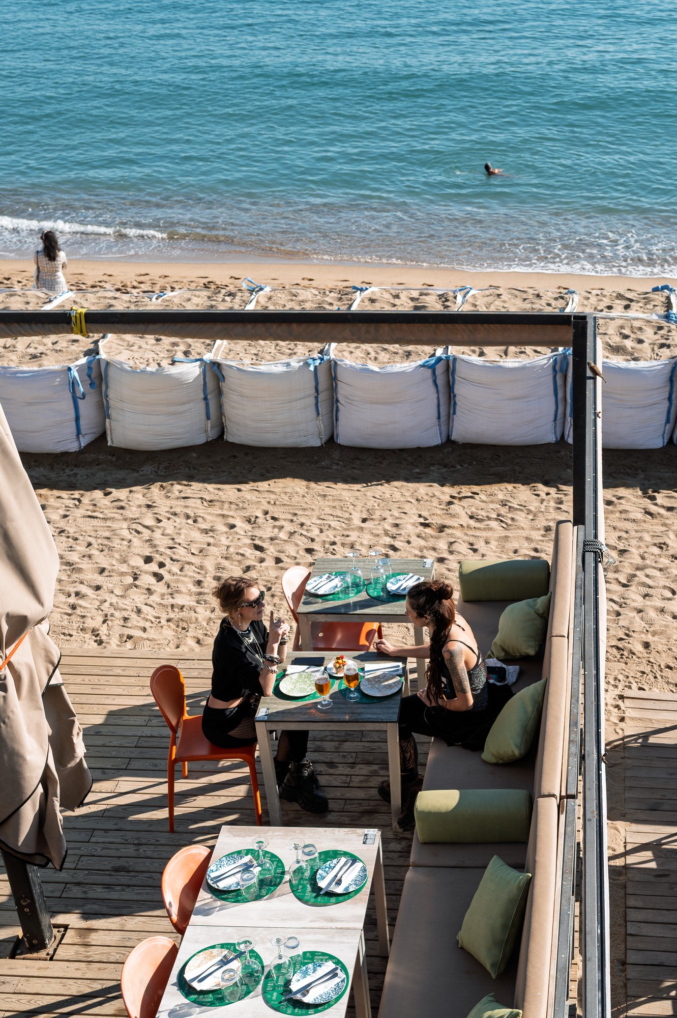 Two women at a beach-bar terrace seen from above, sandbags and sea behind, Barceloneta — Leica Q3