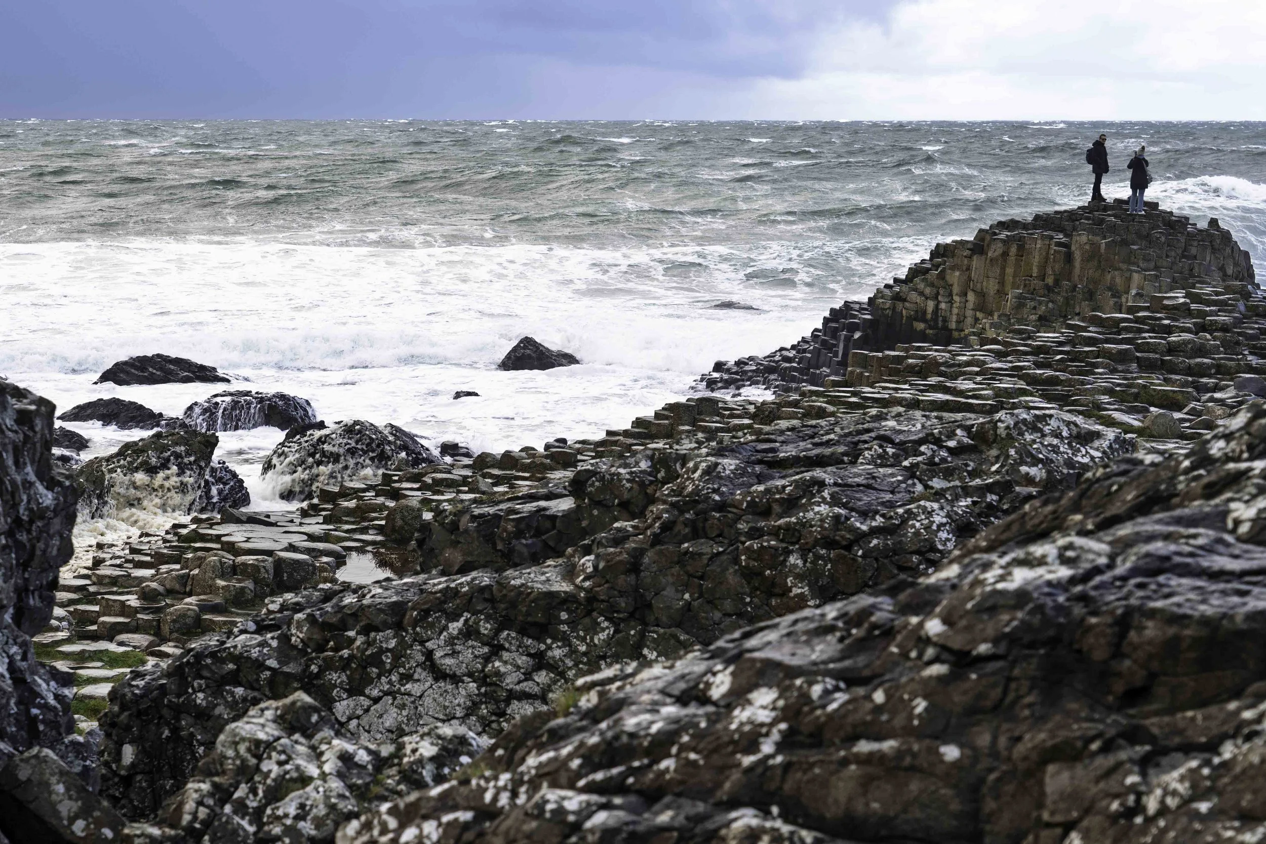Two visitors standing on basalt columns at Giant's Causeway, stormy Atlantic Ocean and dramatic sky behind them, Northern Ireland – bCLPhoto
