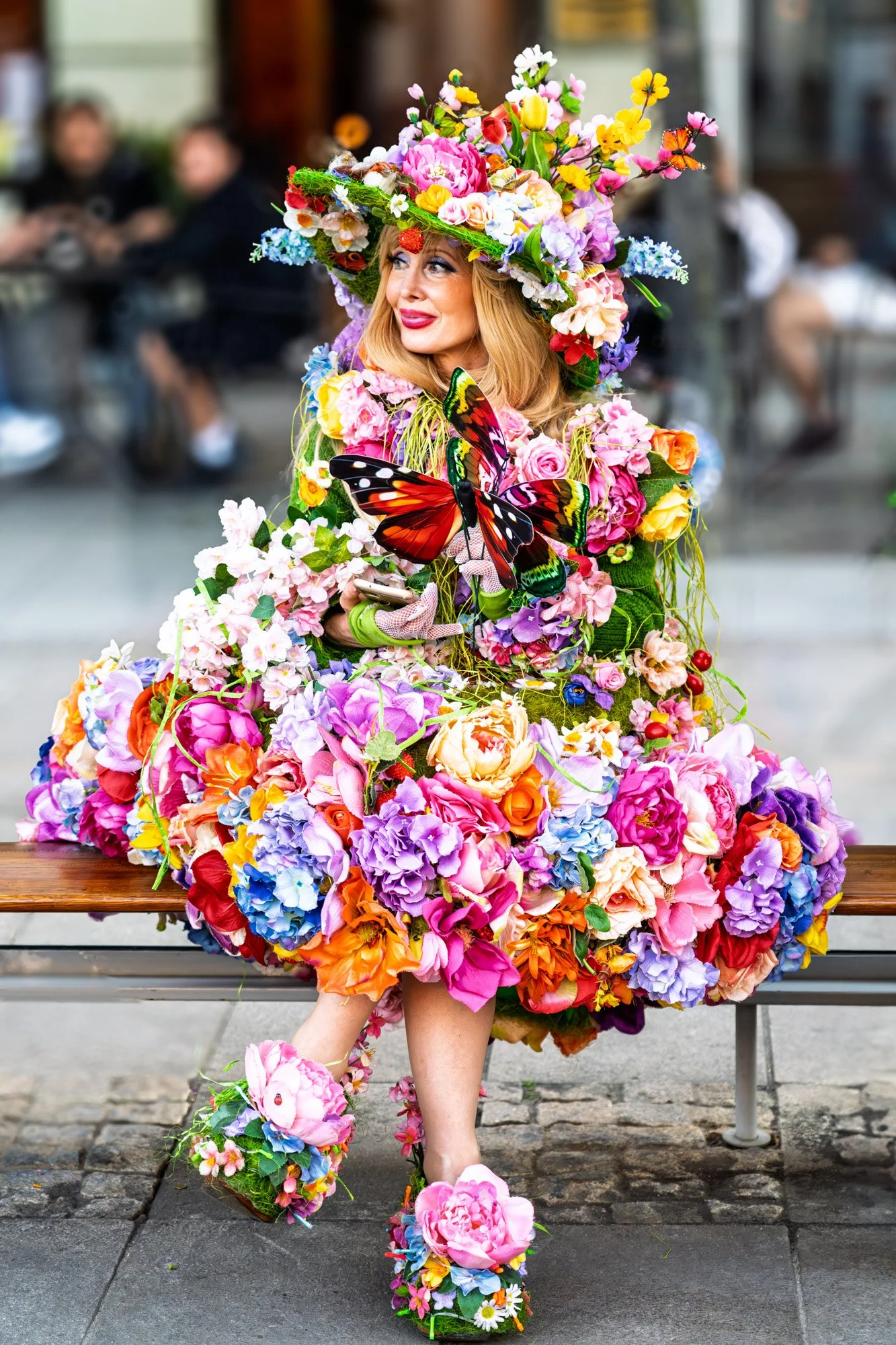 A woman dressed in an elaborate outfit made of colorful flowers, including pink, purple, orange, and yellow, with large butterfly decorations and matching floral shoes, sitting on a bench in an urban outdoor setting.