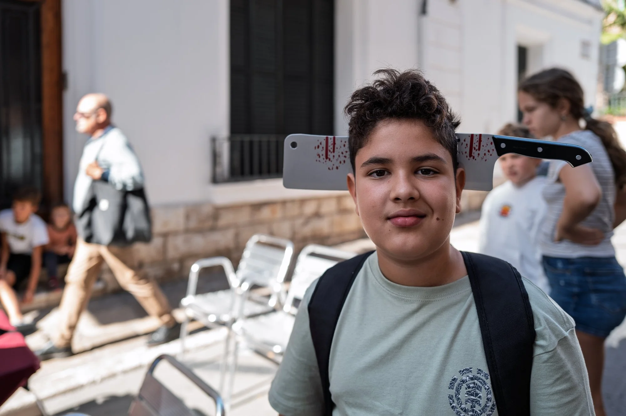 Sitges Zombie Walk: boy with a fake cleaver through his head looks into the lens on a white street — Leica Q3