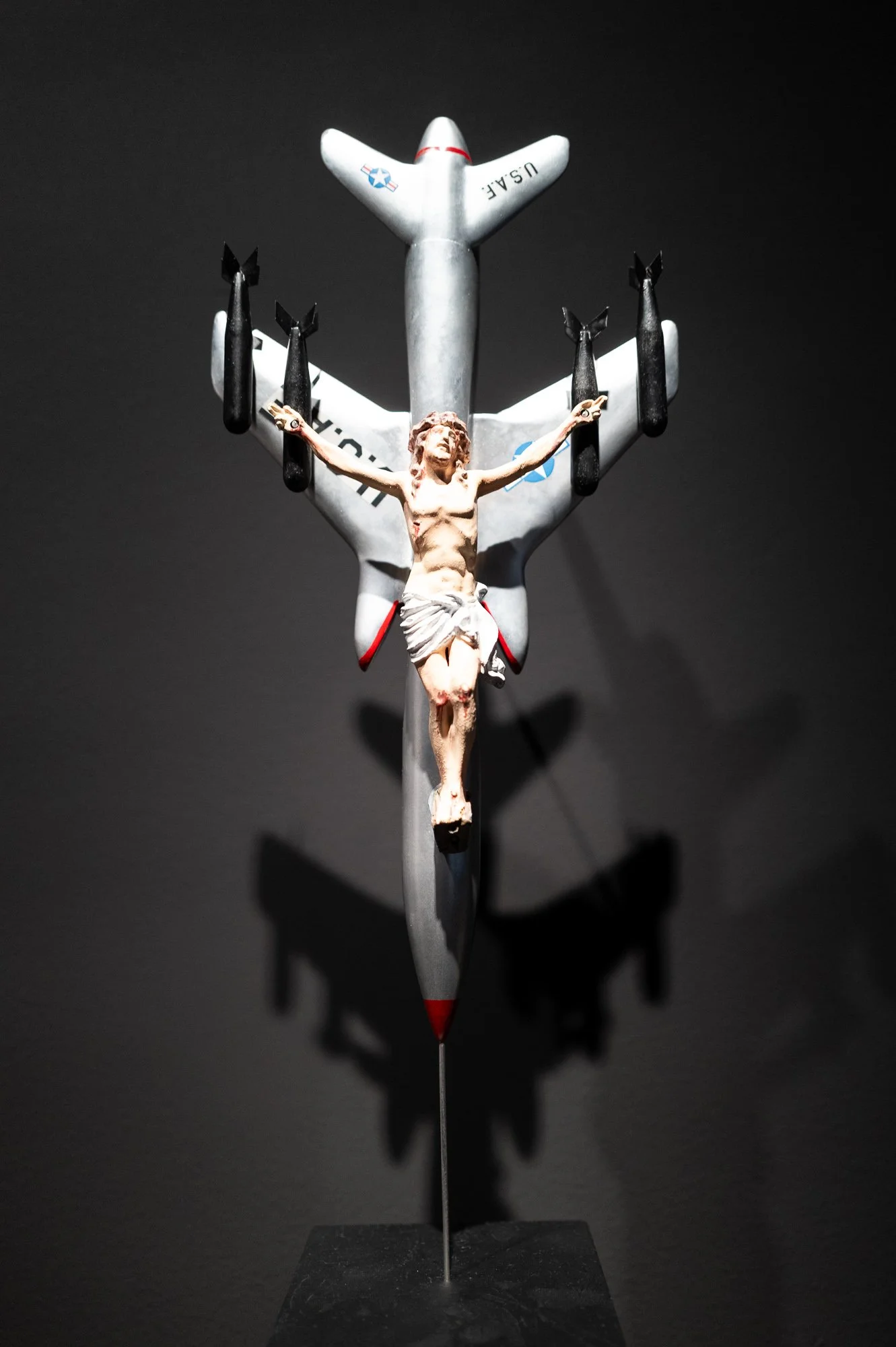 Small figure of Christ crucified on a silver model of a US Air Force bomber carrying black bombs, against a dark wall