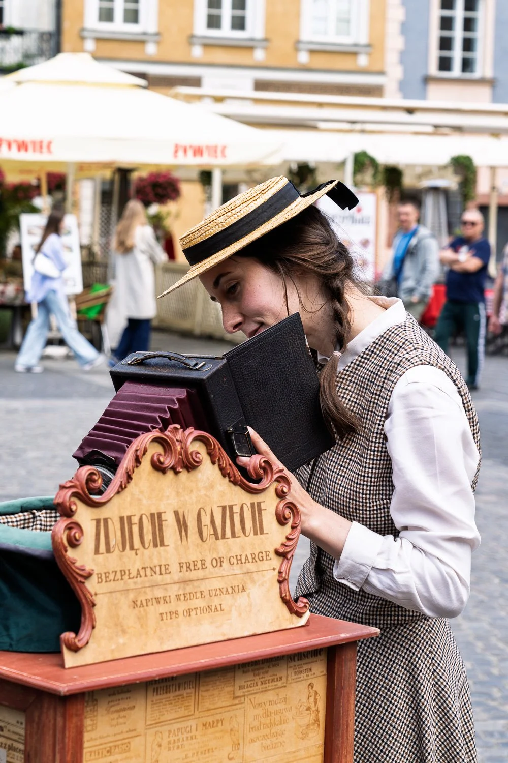 Woman in a straw boater and braids looking into a vintage bellows camera, Old Town Market Square, Warsaw — Sony a7R IV