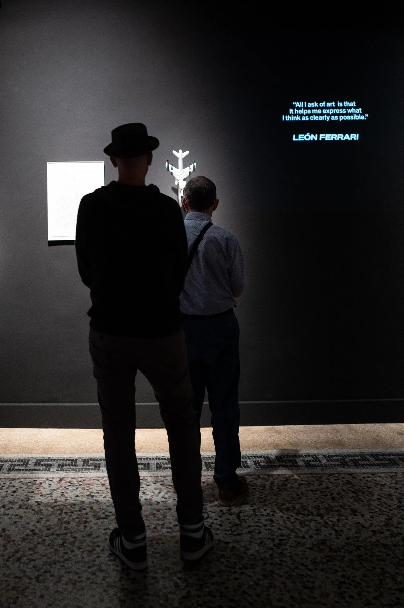 Two men seen from behind look at a small sculpture of a bomber on a dark wall, a quotation projected in blue beside it