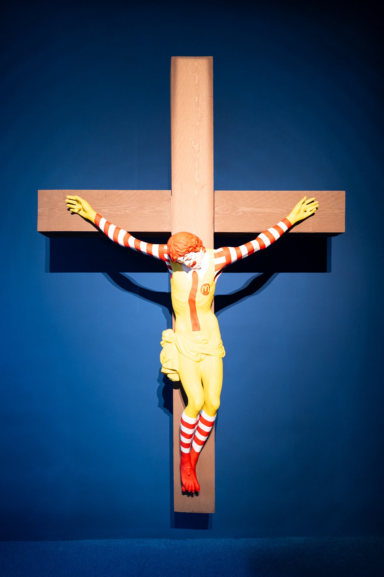 A crucified figure painted in the colours of a fast-food clown on a wooden cross, against a deep blue wall