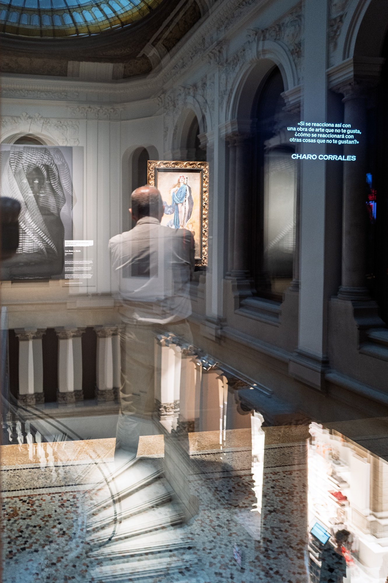A man photographs a framed painting of the Virgin, layered in a glass reflection with the stairwell, a quote projected on the wall