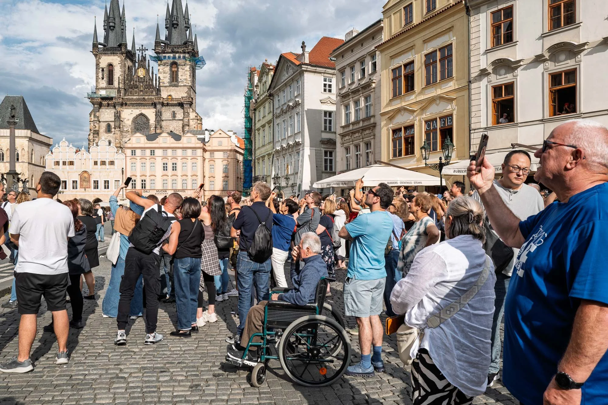 Crowd raising phones before Týn Church, a man in a wheelchair among them, Old Town Square, Prague — Leica Q3