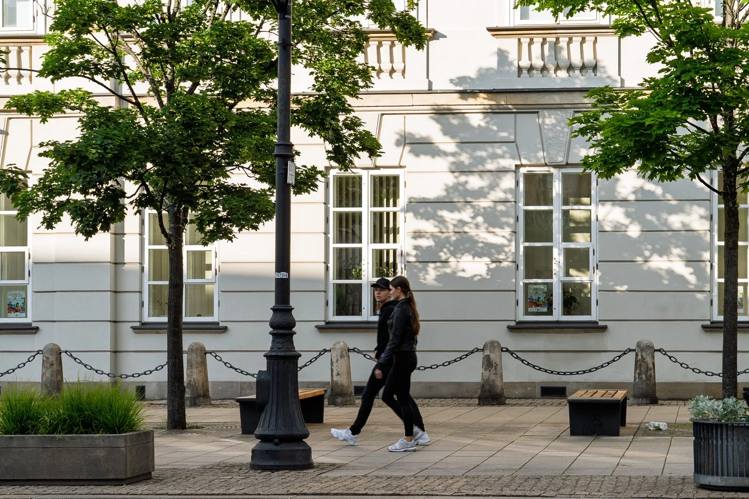 Couple in black walking past tree shadows on a pale façade and chained bollards, Krakowskie Przedmieście — Sony a7R IV