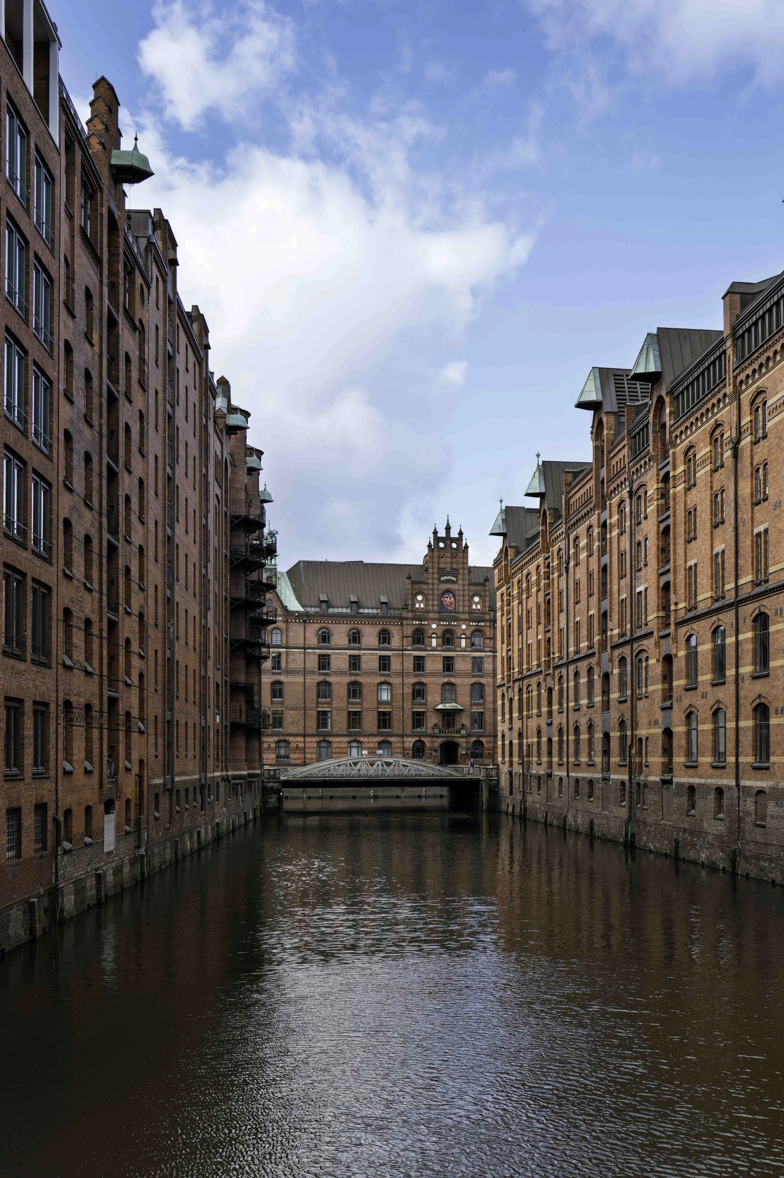 Narrow Speicherstadt fleet canal flanked by red-brick warehouses, small iron bridge and clock tower in background, Hamburg — Leica Q3