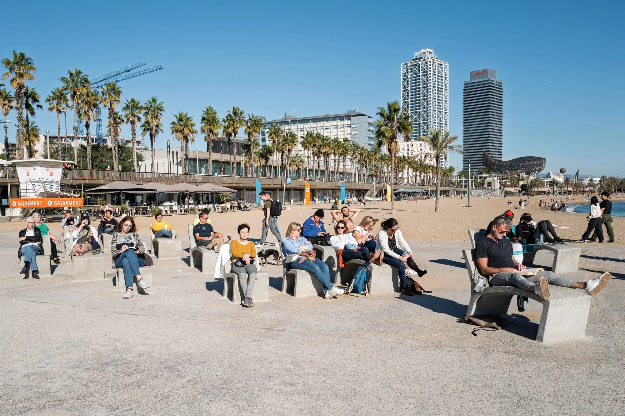 People reading on concrete loungers, Port Olímpic towers behind, Barceloneta beach — Leica Q3