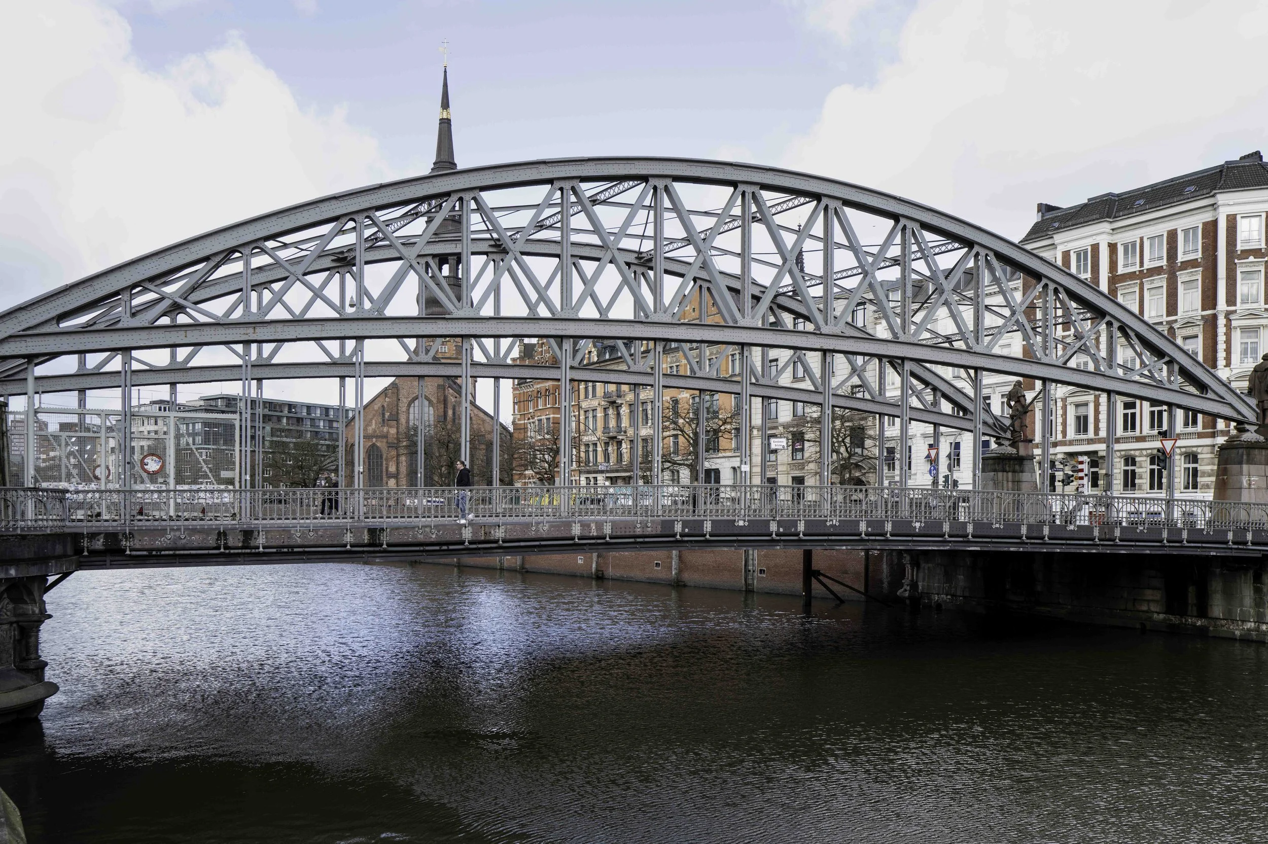 Wide steel truss arch bridge over Hamburg canal with Gothic church spire visible behind — Leica Q3