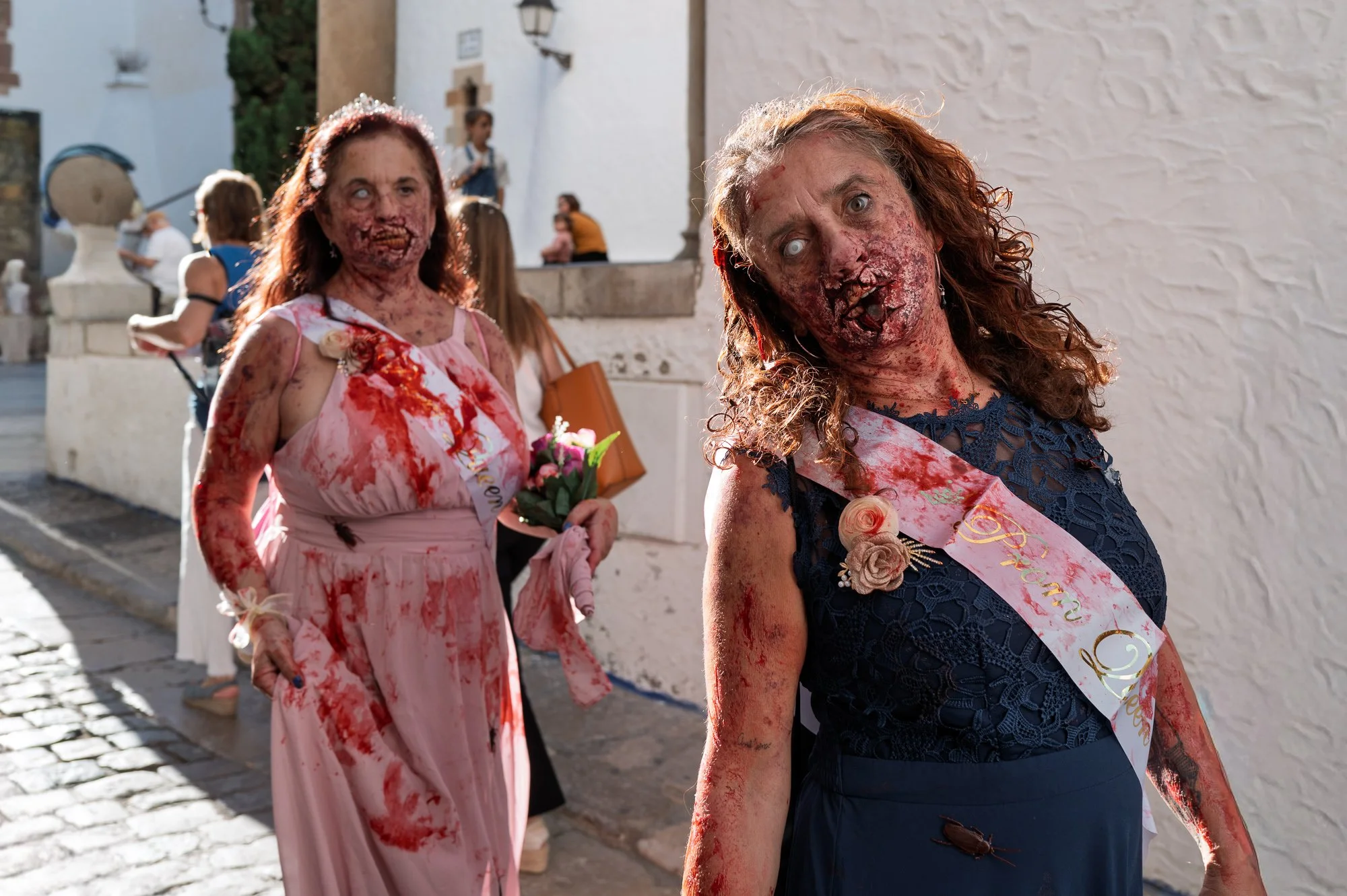 Two zombie bridesmaids with satin sashes and white eyes on a cobbled lane in Sitges — Leica Q3