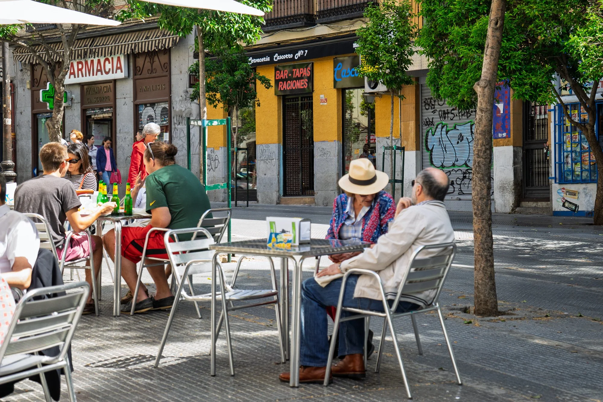 People at metal café tables in Plaza de Cascorro, a woman in a straw hat facing a man, a pharmacy and bar behind