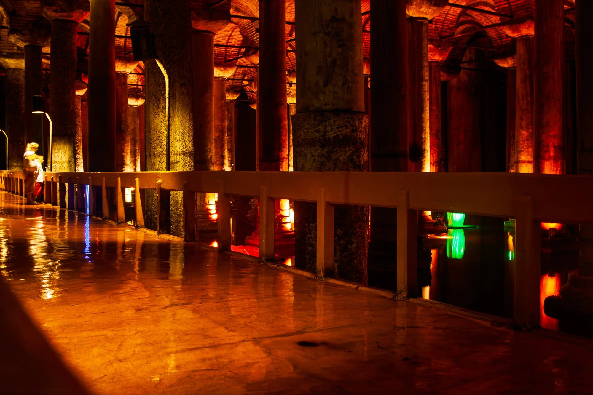 Wet walkway reflecting orange light beside a low white railing, green and red lights in the water, Basilica Cistern