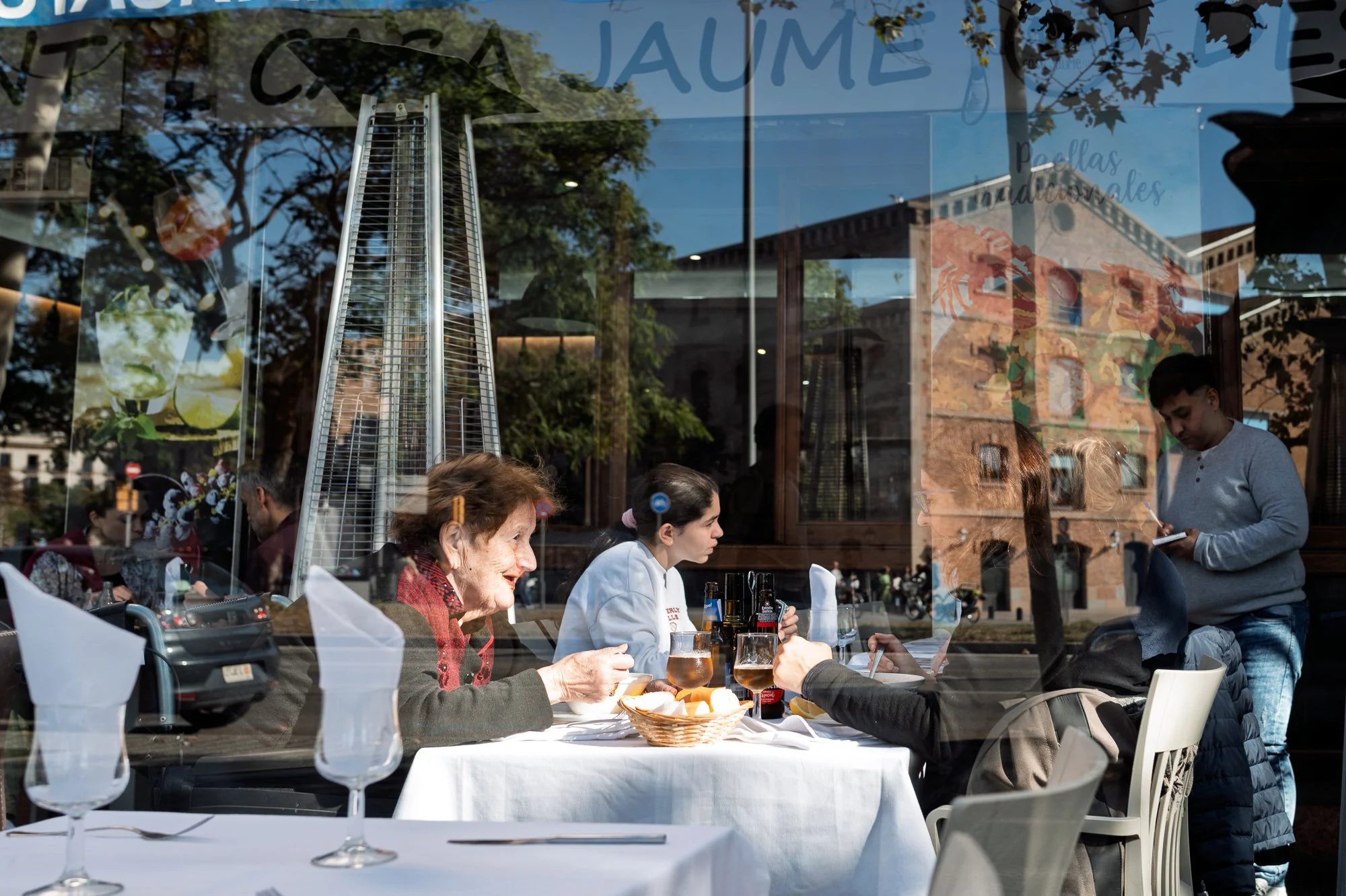 Diners behind a restaurant window reflecting a brick building, Joan de Borbó, Barceloneta — Leica Q3