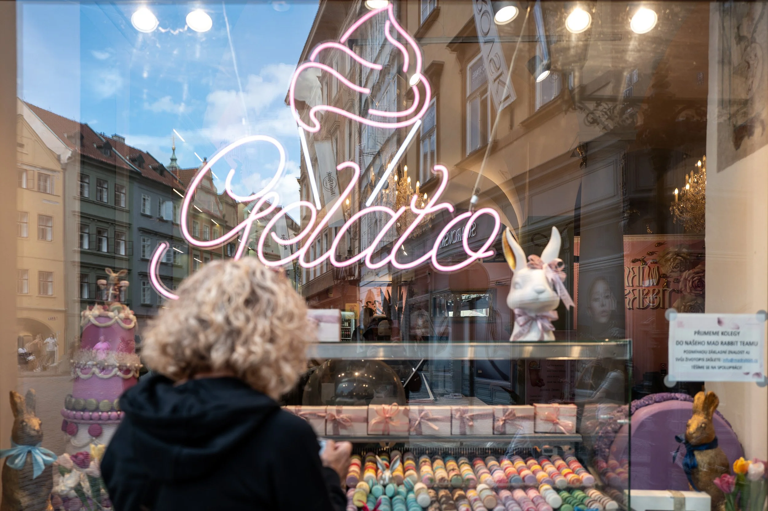 Woman at a pastry shop window, pink neon Gelato sign and Old Town façades reflected, Prague — Leica Q3