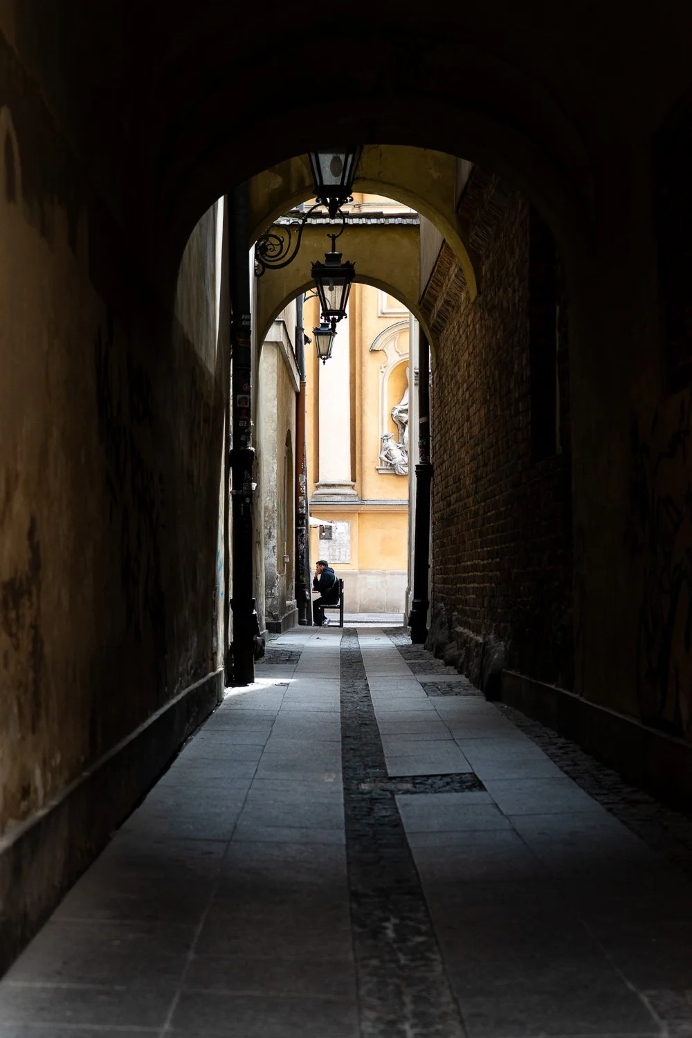 Warsaw Old Town street photography: a man sits on a bench at the end of a dark arched passage with lanterns — Sony a7R IV