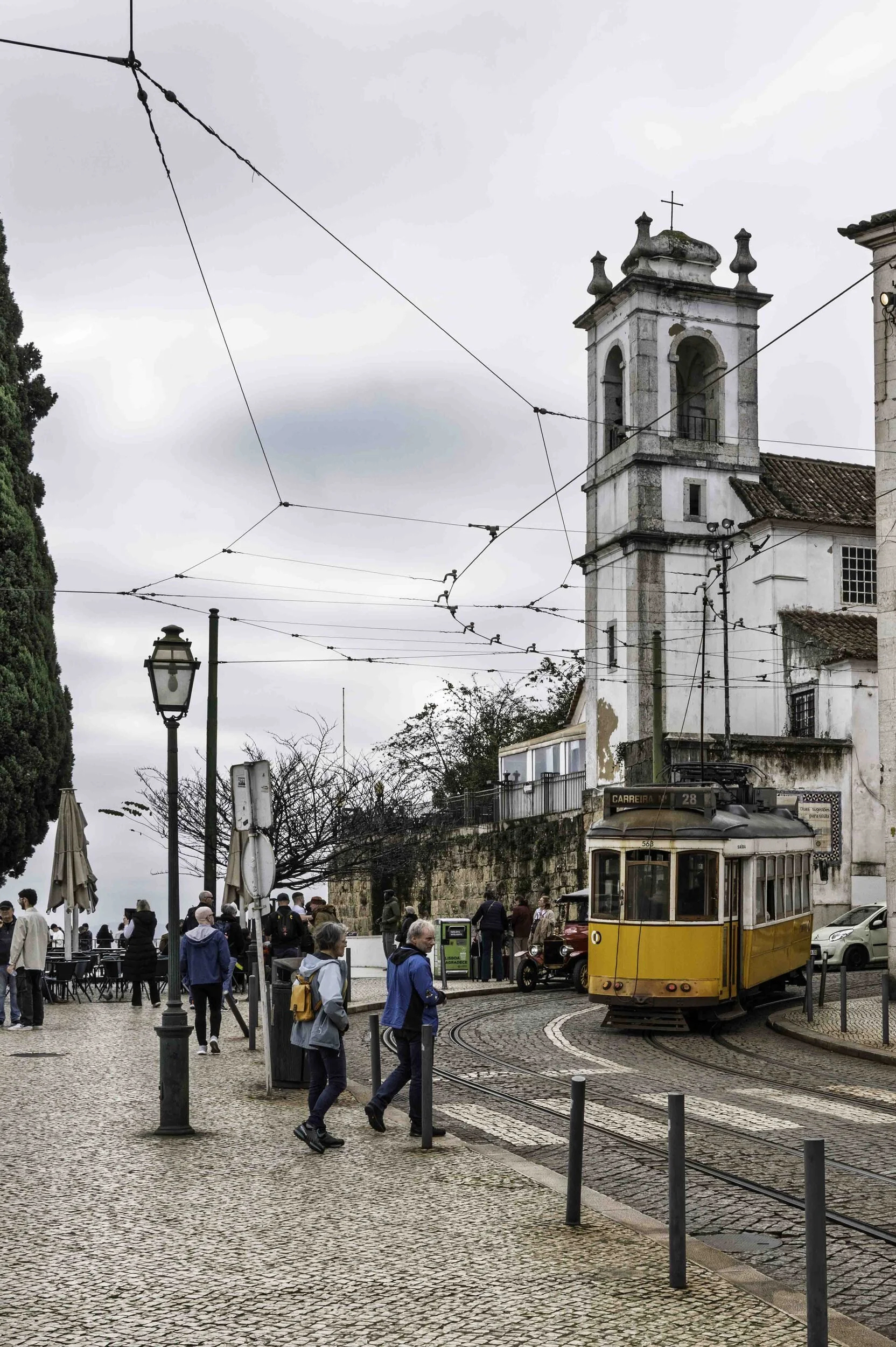 Yellow tram 28 rounding a corner in Alfama, Lisbon street photography with church tower — Leica Q3