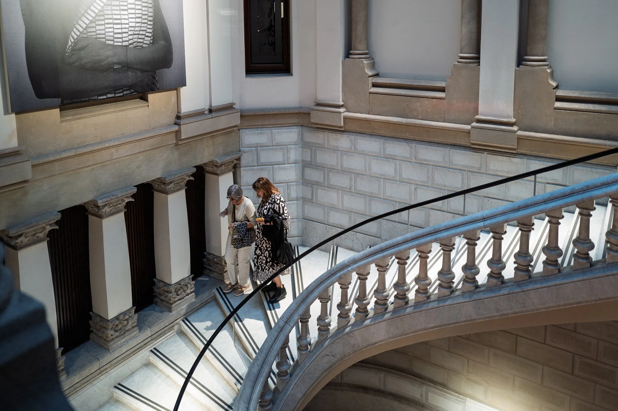 Two women walk down a curving marble staircase together, one in a black and white patterned dress, past stone pilasters